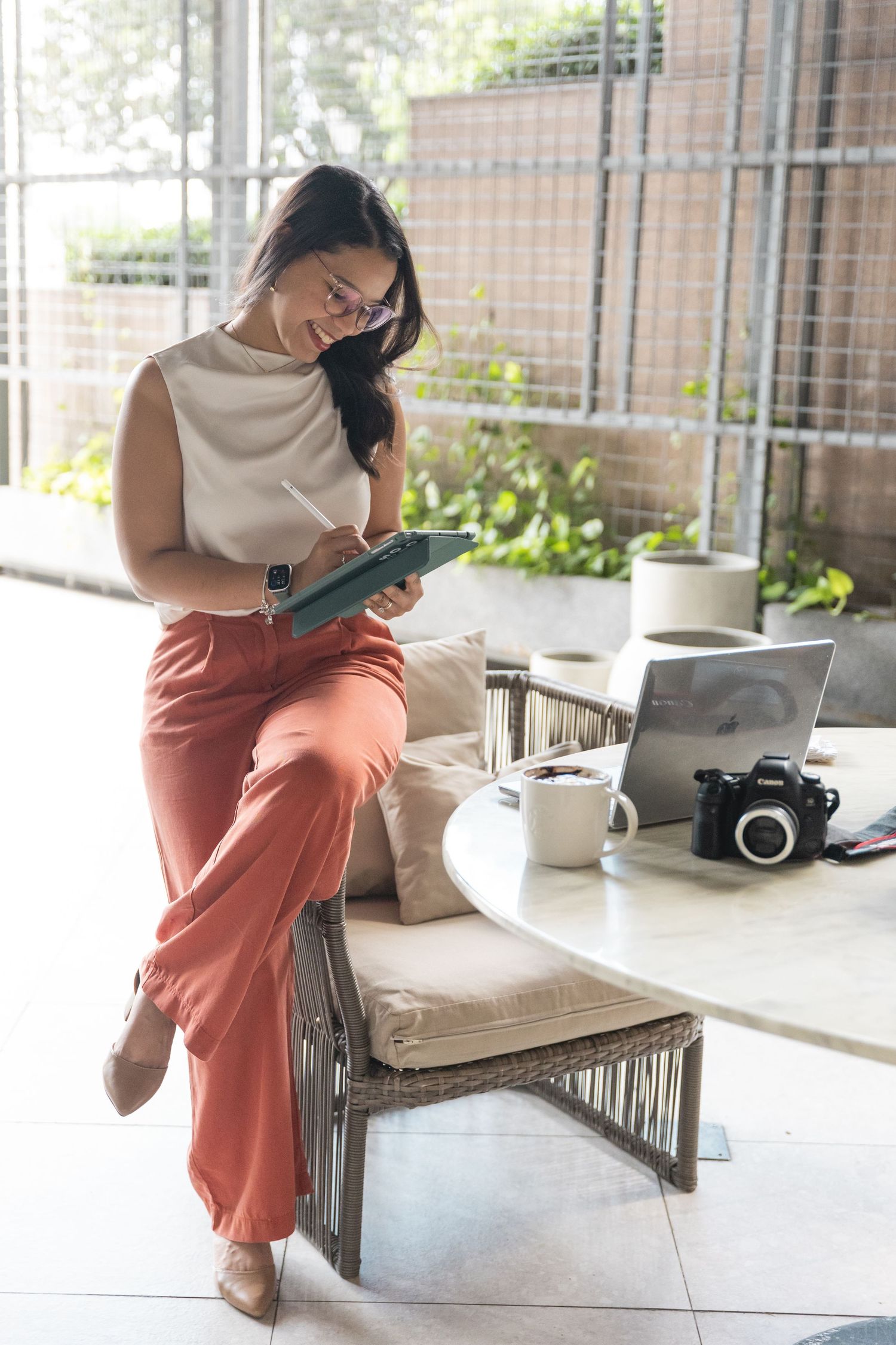 A stylish woman in orange trousers writes in a notebook beside a laptop and camera at a bright cafe.