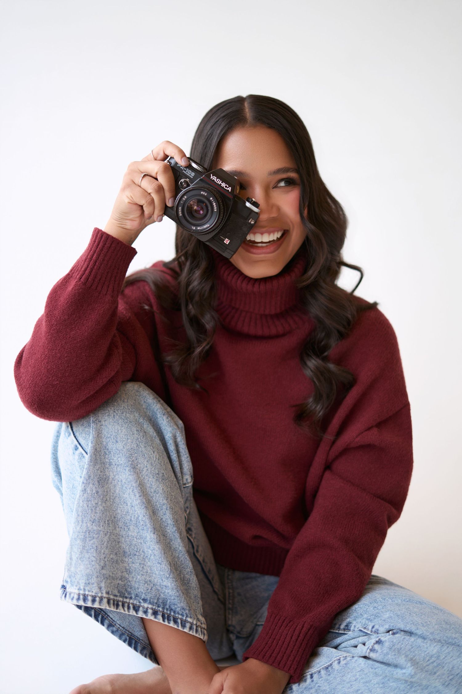 Smiling young woman in burgundy turtleneck sweater and jeans holding a vintage film camera up to her eye.