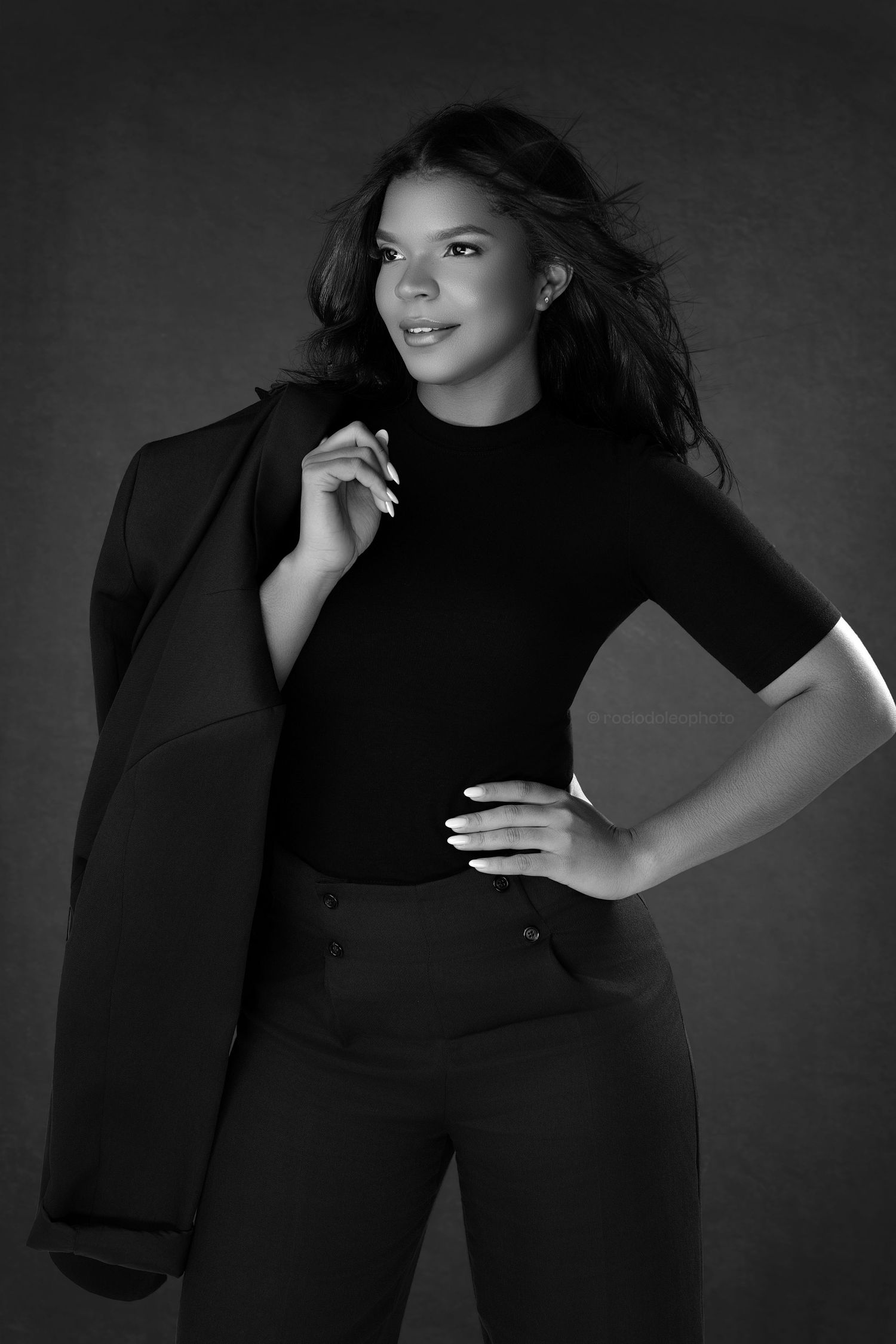 Black and white portrait of a young woman in a black turtleneck and blazer, posing confidently with hand on hip.
