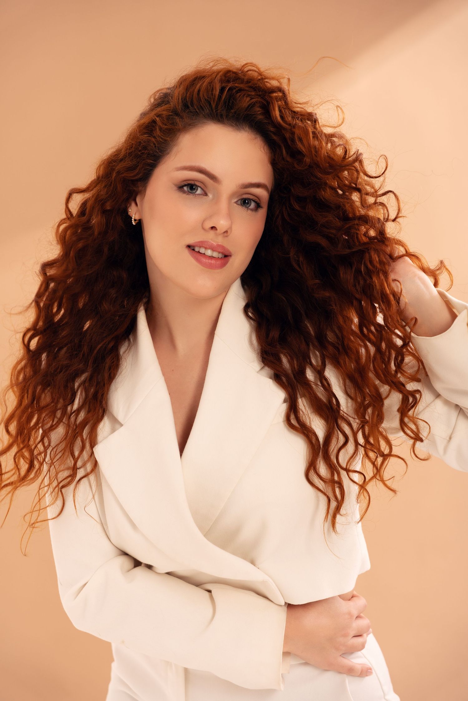 Professional fashion portrait featuring a white blazer and flowing curly red hair against a peach backdrop.