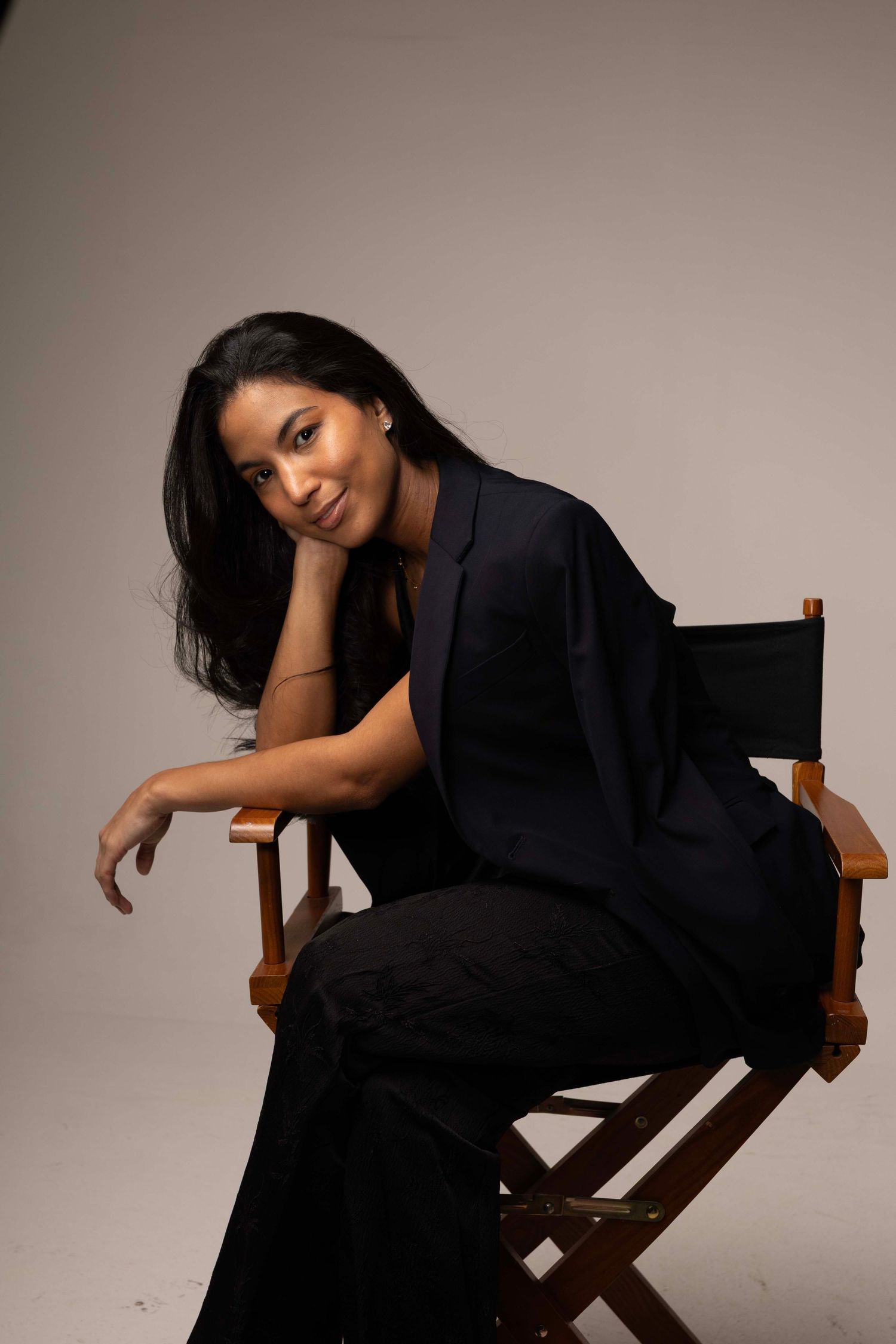 A young woman in black outfit sits gracefully in a director's chair, smiling against a neutral gray background.