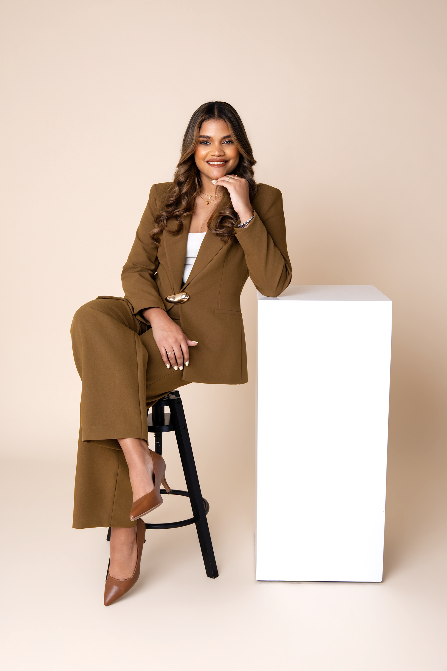 Woman in brown suit sitting on stool next to white pedestal, smiling confidently in a professional studio setting.
