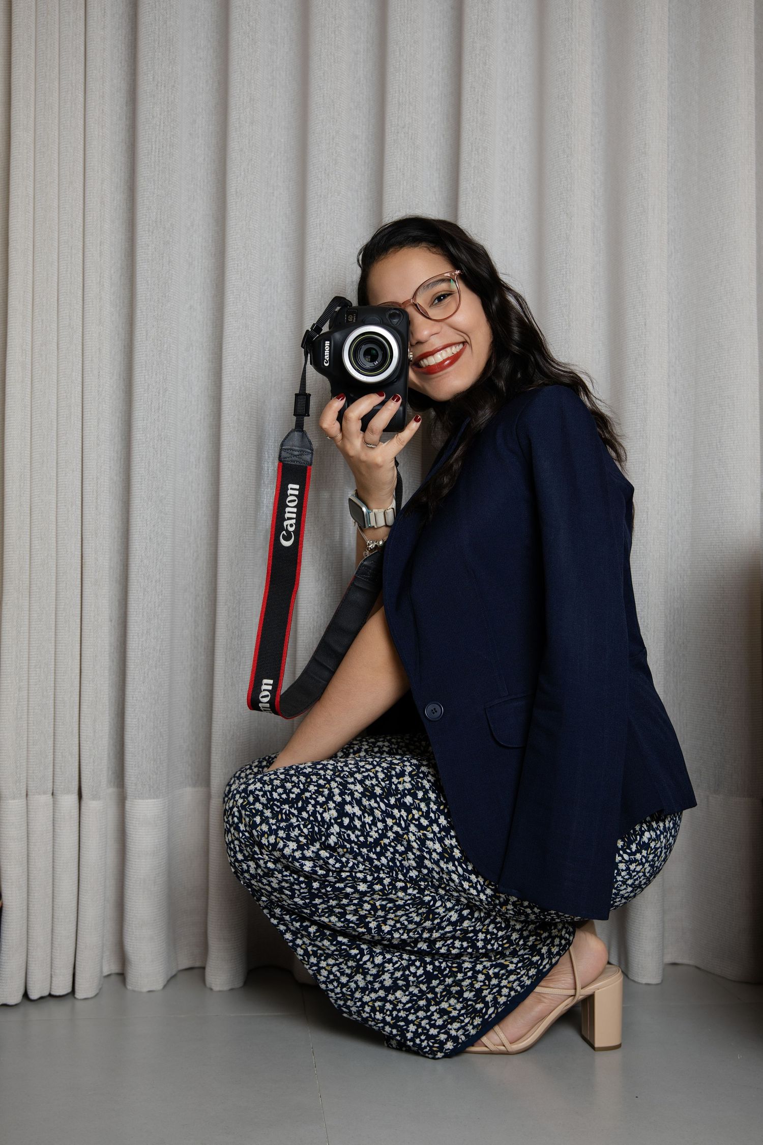 Smiling woman in navy blazer and floral skirt crouching while holding a Canon DSLR camera near a white curtain.