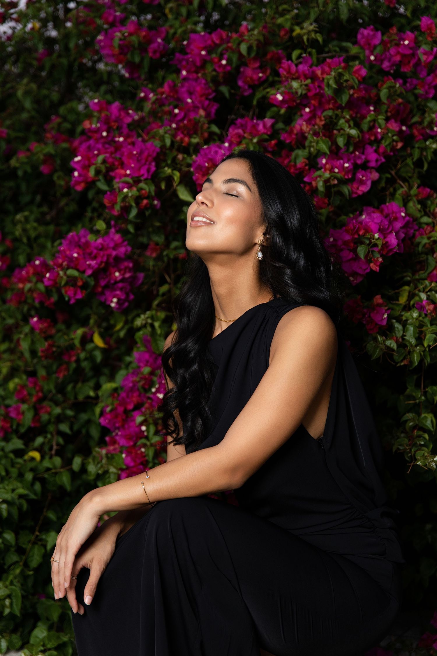 A figure in a black dress poses expressively against a backdrop of vibrant magenta bougainvillea flowers in a garden setting.