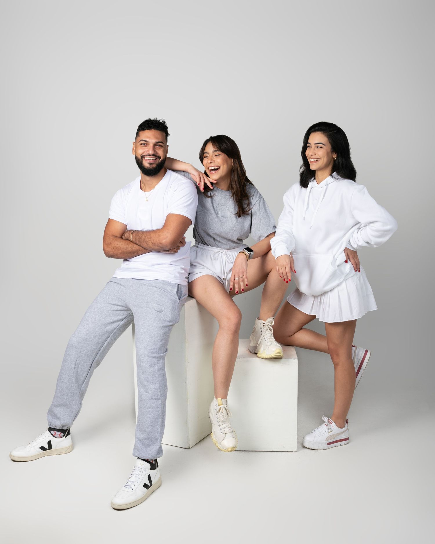 Three people in casual white and grey outfits pose playfully against studio backdrop.