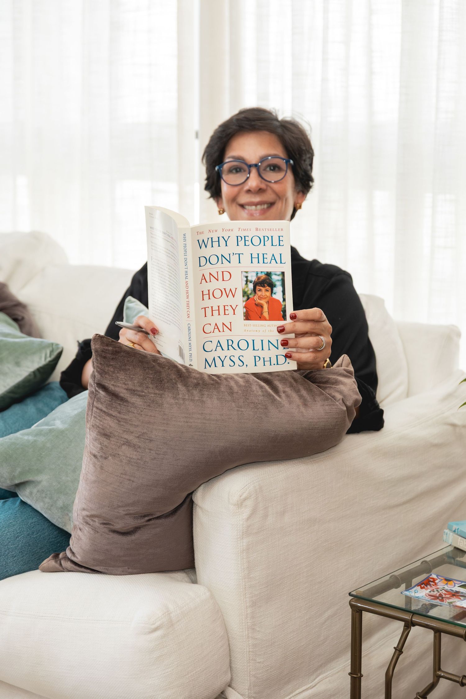 Woman smiling on white sofa holding Why People Dont Heal And How They Can by Caroline Myss PhD book.