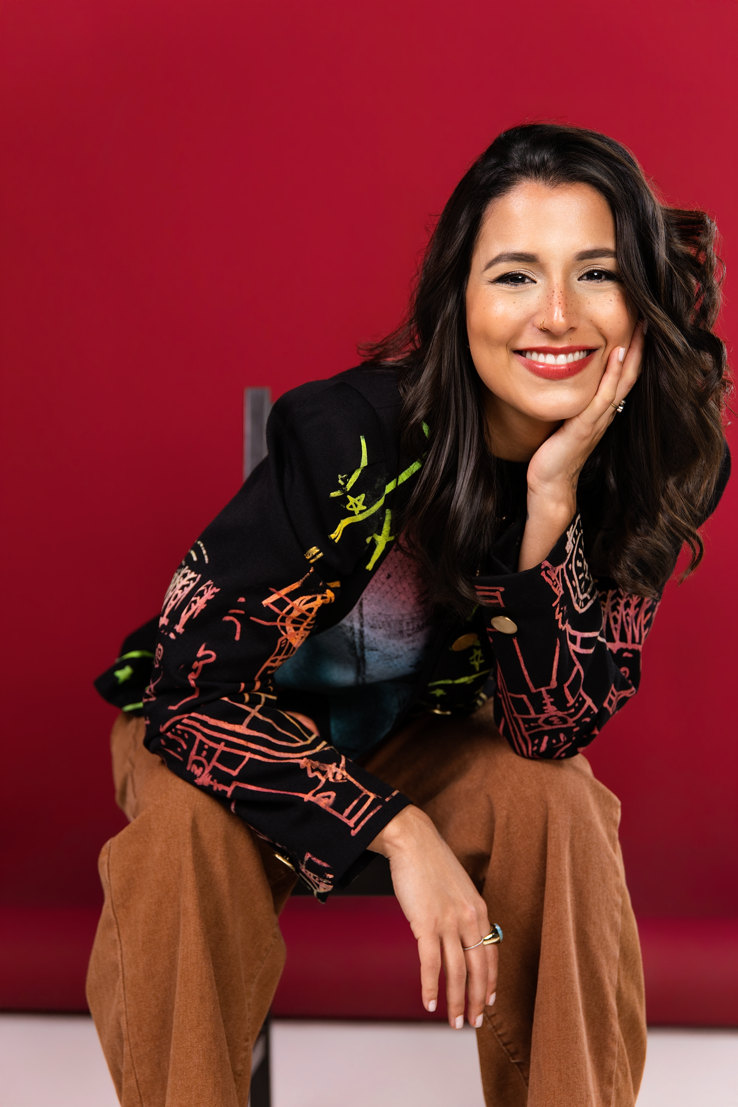 Smiling woman in colorful printed blazer and tan pants poses seated against a bold red background.