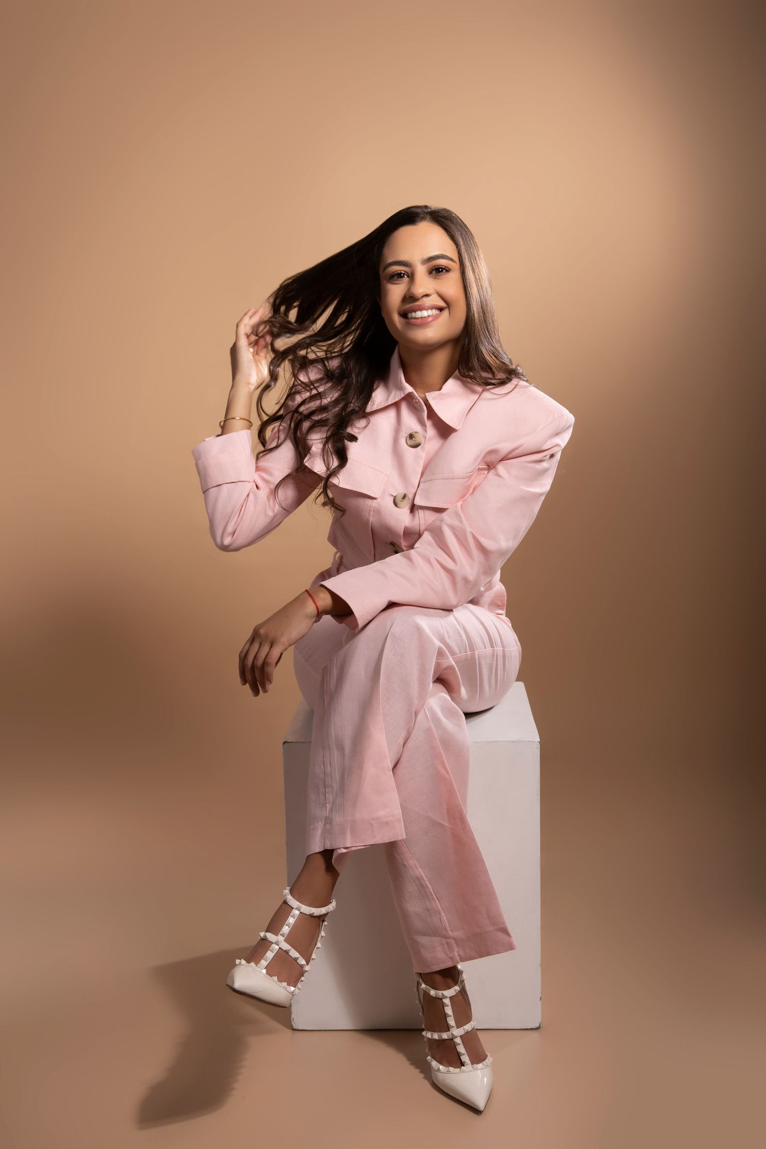 Joyful portrait of person in pink suit sitting on white cube with flowing hair.