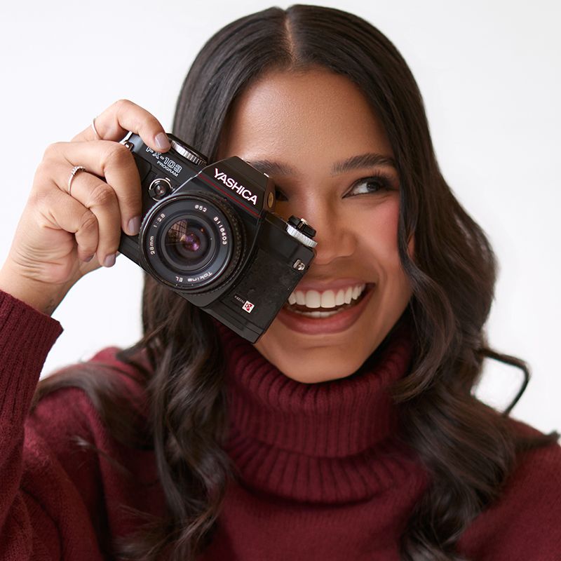 Smiling young woman holding a Yashica film camera, wearing a burgundy turtleneck sweater against a white background.