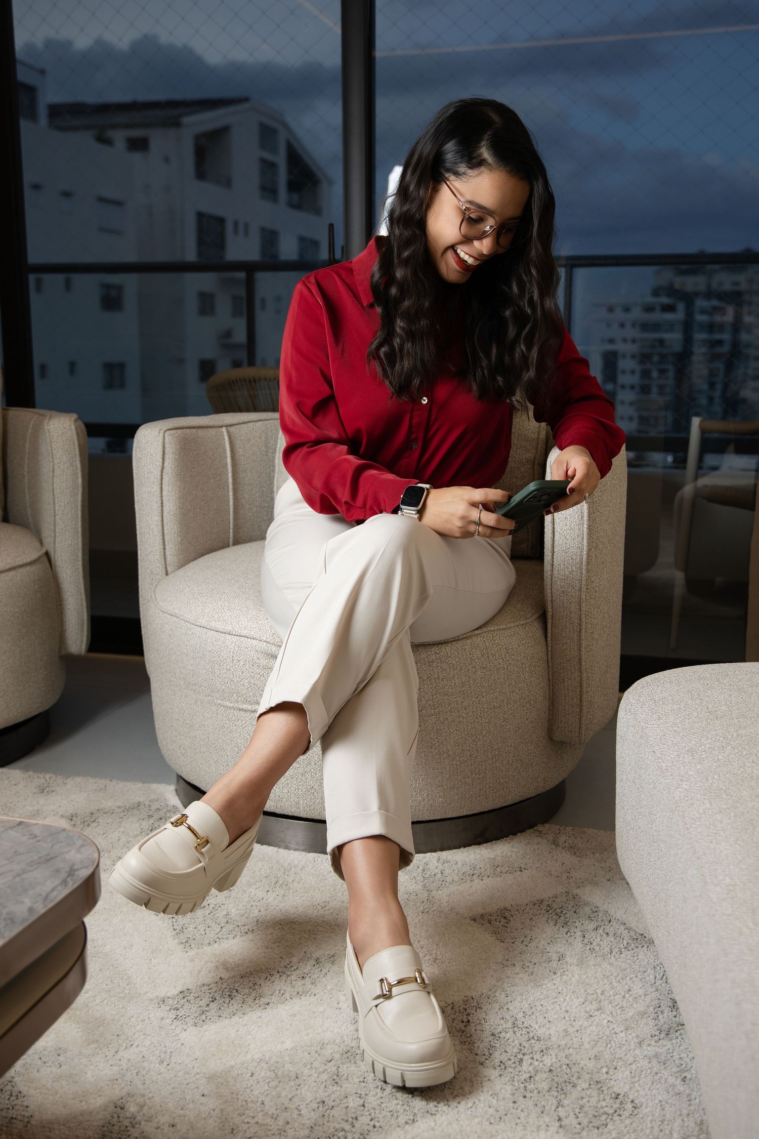 Young woman in red shirt and white pants sits in beige armchair smiling at her smartphone, wearing cream loafers.