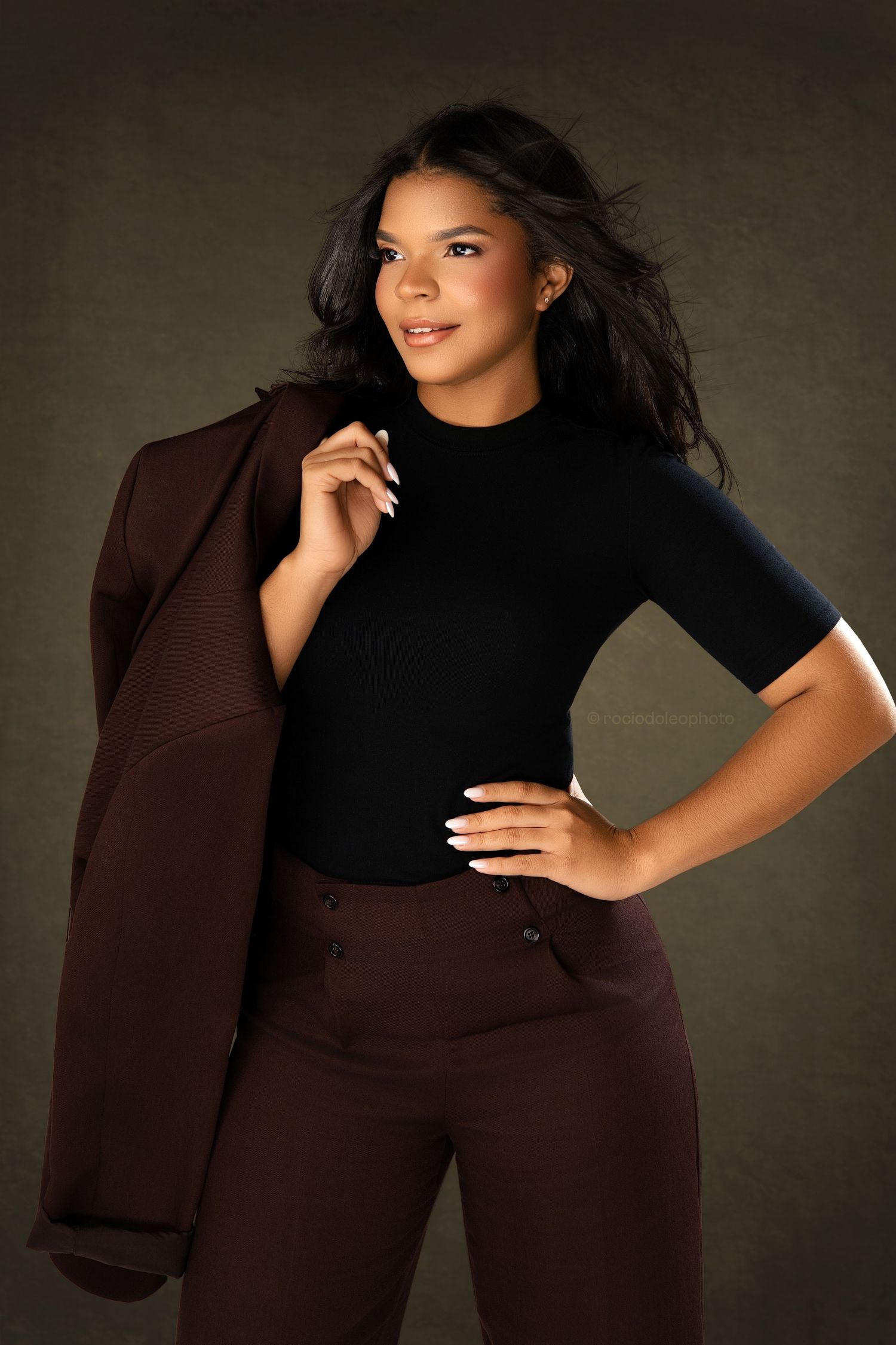Young woman in black turtleneck and brown trousers poses confidently holding a matching blazer over her shoulder.