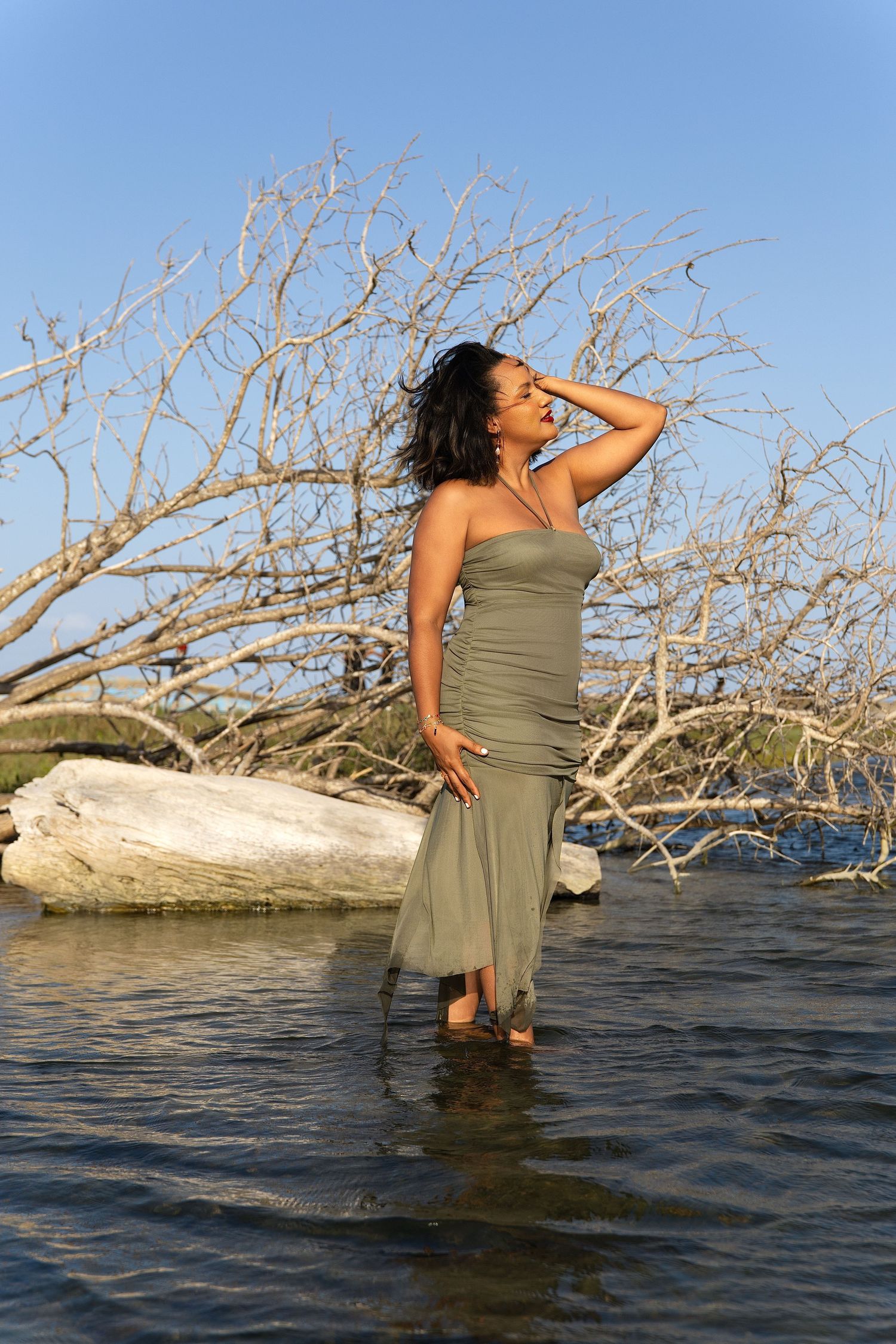Woman in olive strapless maxi dress standing in shallow water among driftwood branches under clear blue sky.
