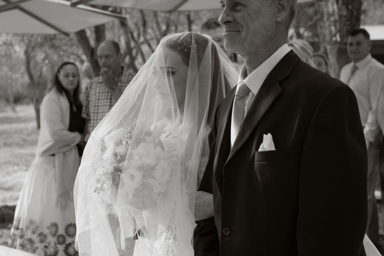 A bride in a white veil walks with an older man in a black suit during an outdoor wedding ceremony.