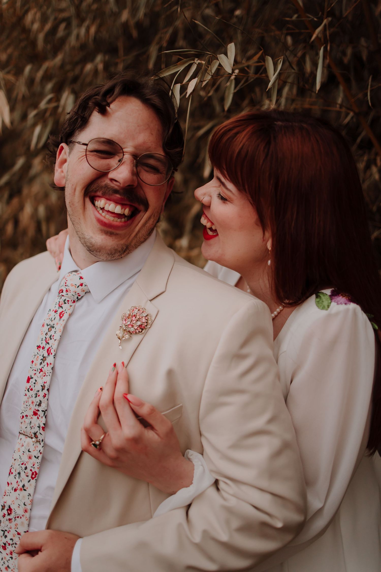 Joyful newlywed couple laughing together outdoors, groom in cream suit with floral tie, bride in white dress.