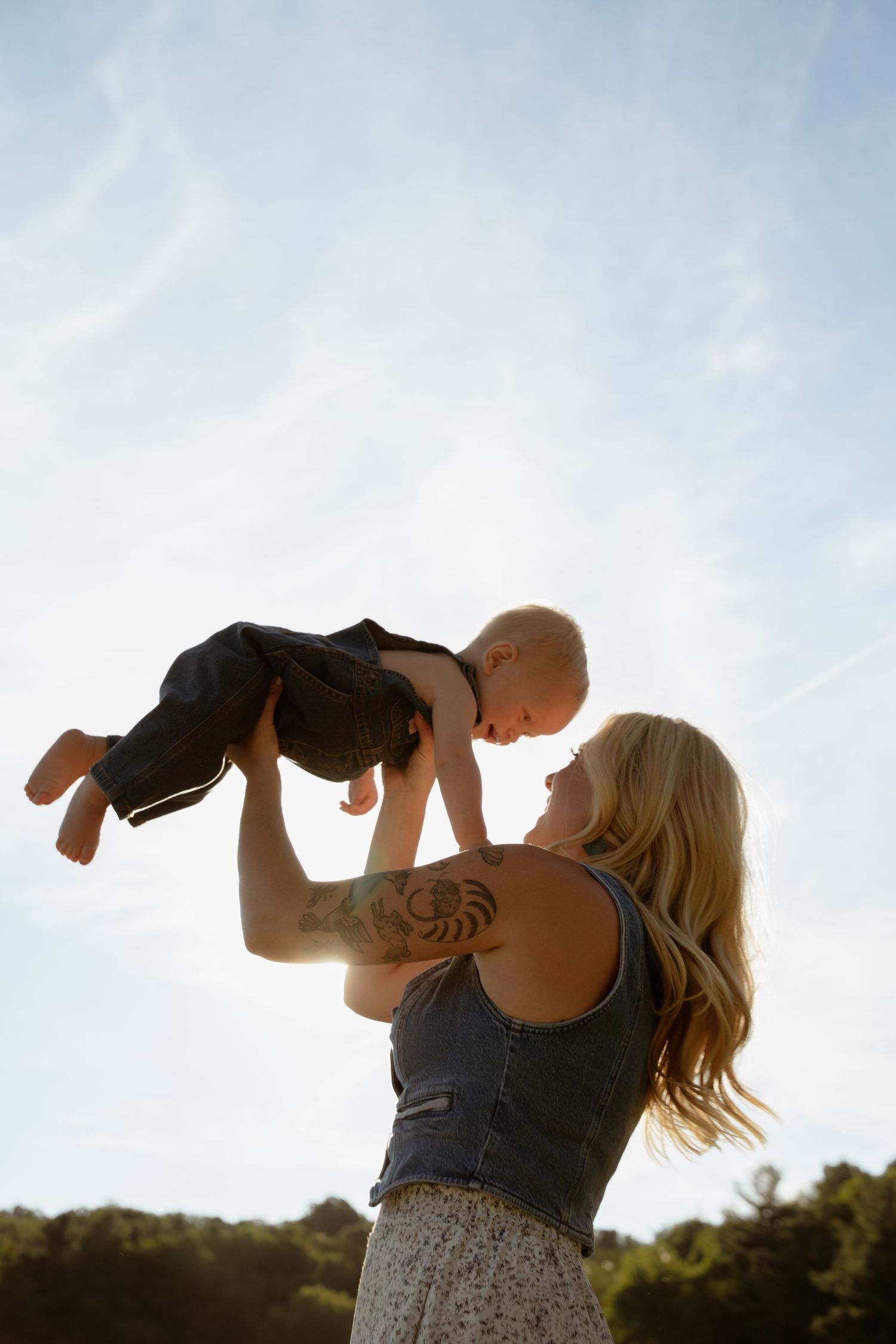 Silhouetted mom playfully lifting child against bright blue sky background.