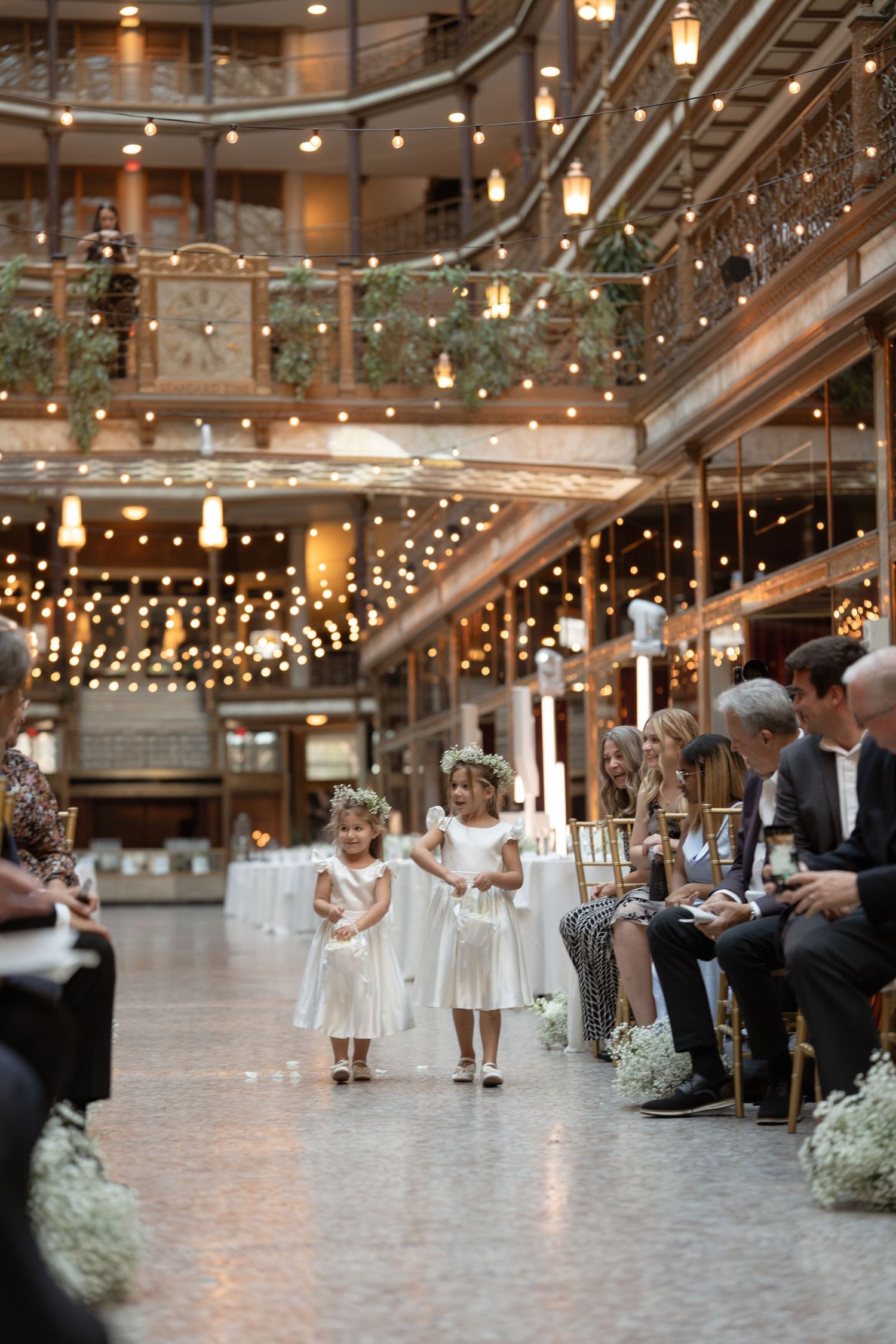 Young flower girls in white dresses dance and twirl under twinkling string lights at an elegant indoor wedding reception.