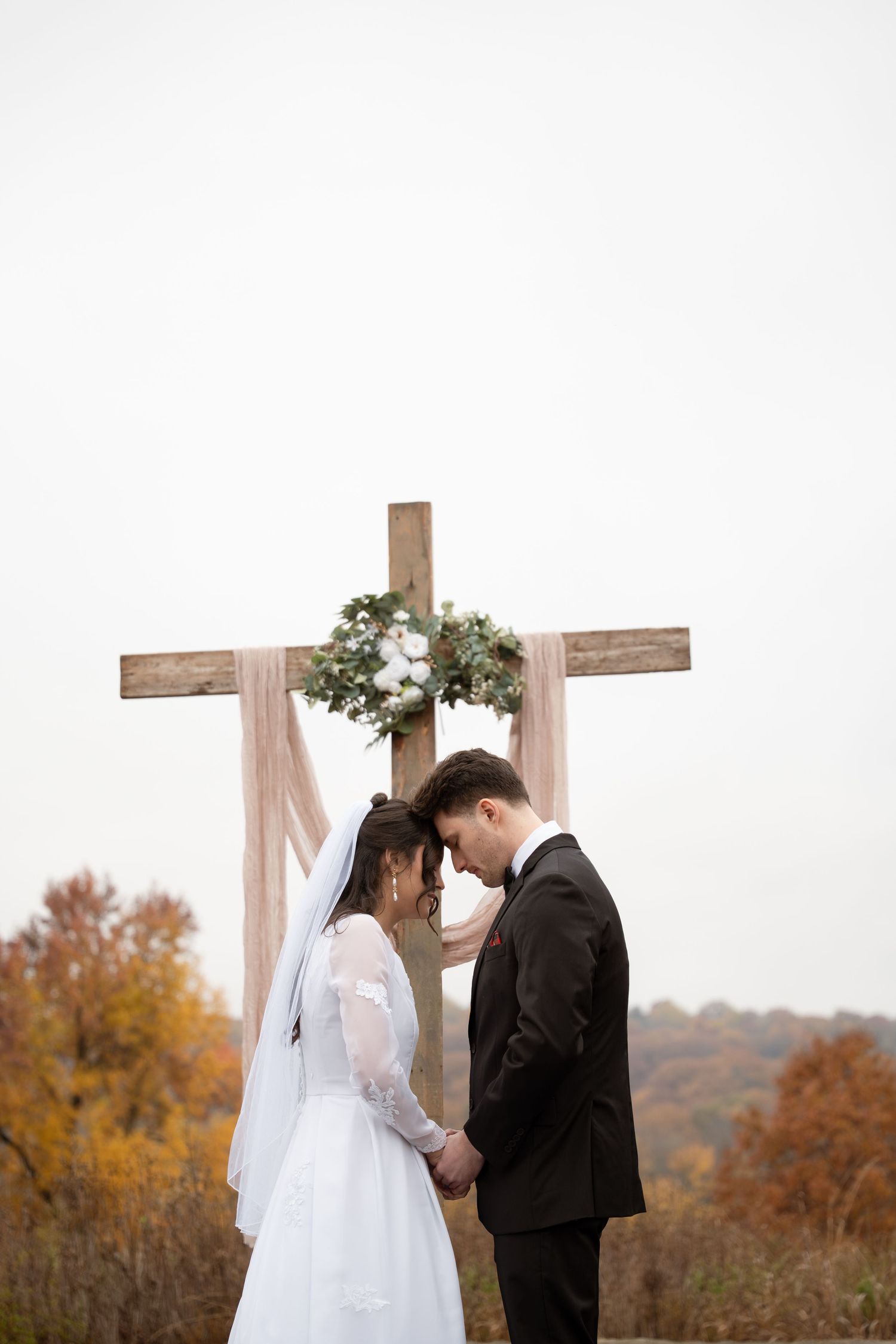 An intimate wedding ceremony moment under a wooden cross decorated with greenery overlooking a vast valley of fall foliage.