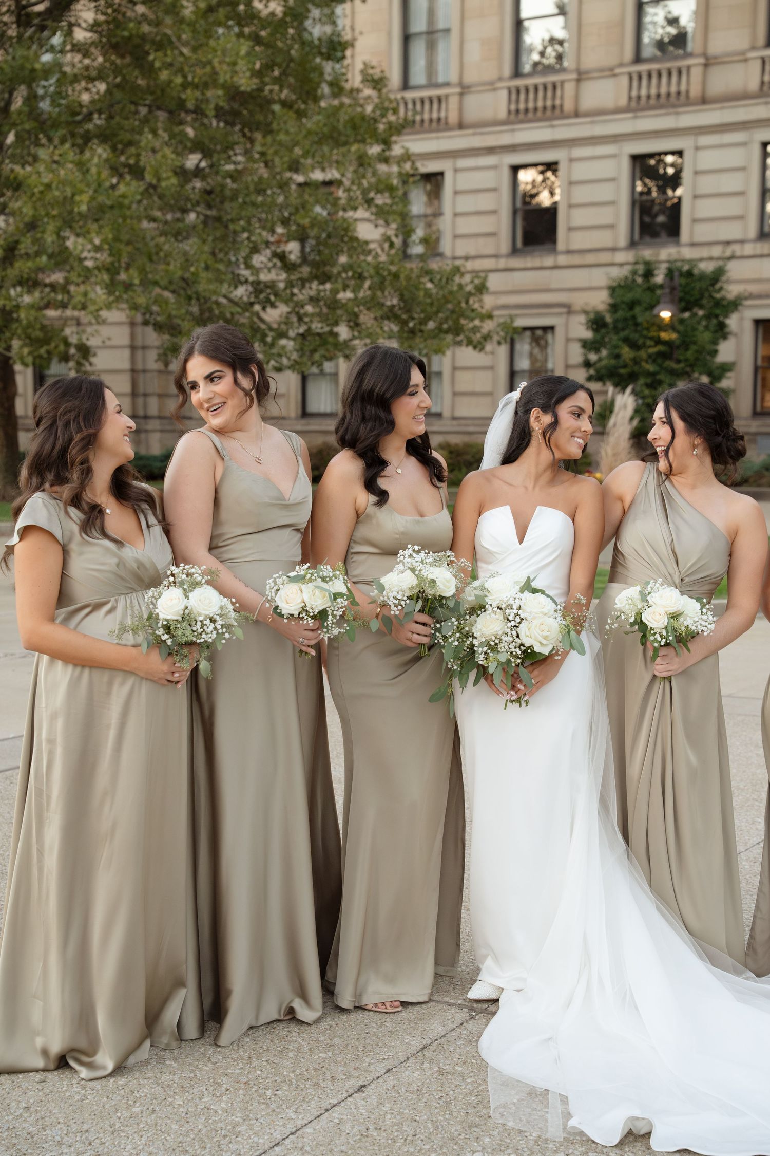 A bridal party in neutral beige bridesmaid dresses and white wedding gown pose together outdoors by a stone building.