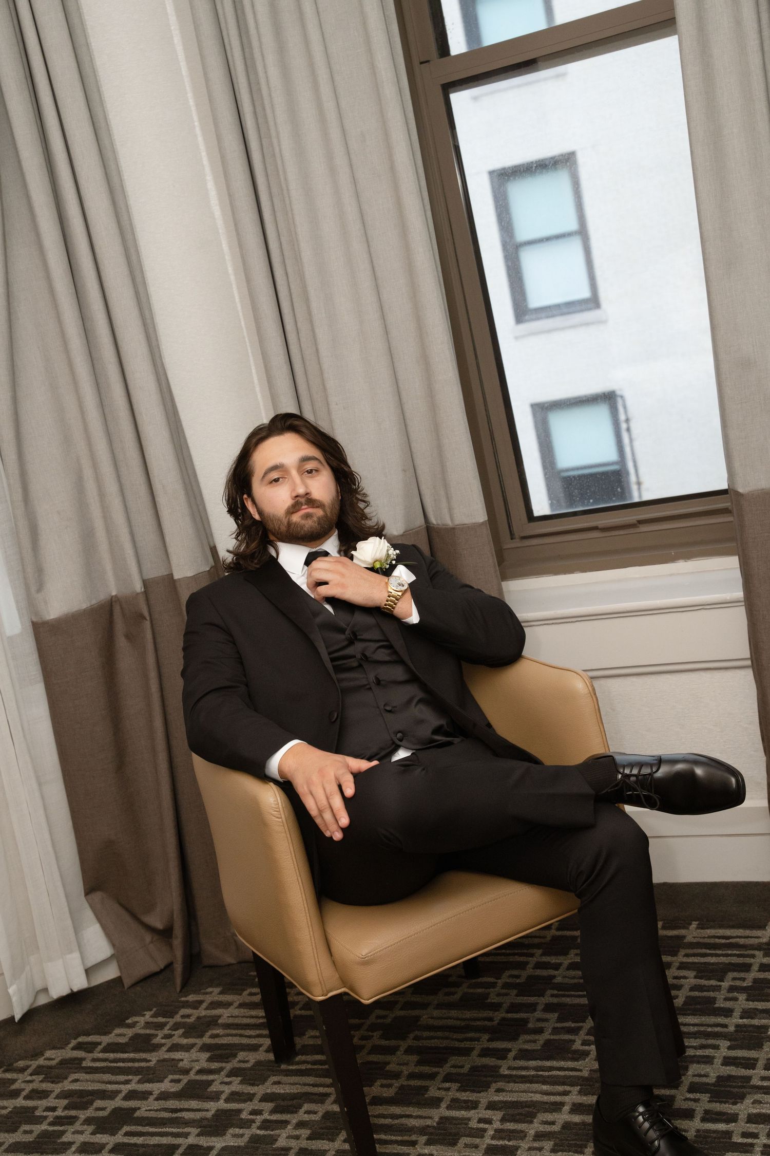 Groom in black suit lounges dramatically on tan chair in hotel room.