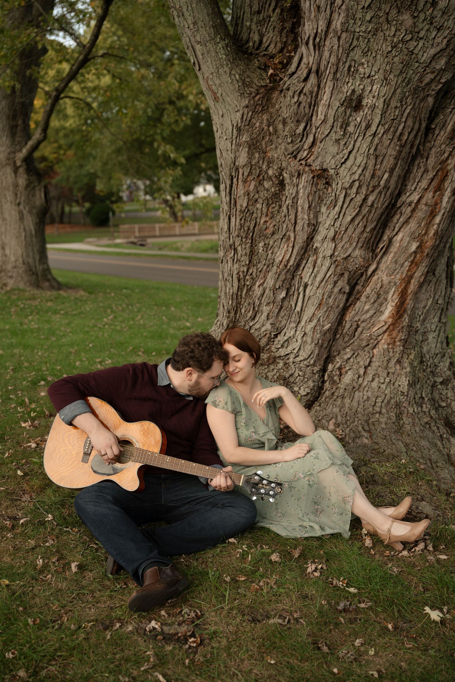 Couple relaxes under large tree trunk while playing acoustic guitar in park setting.