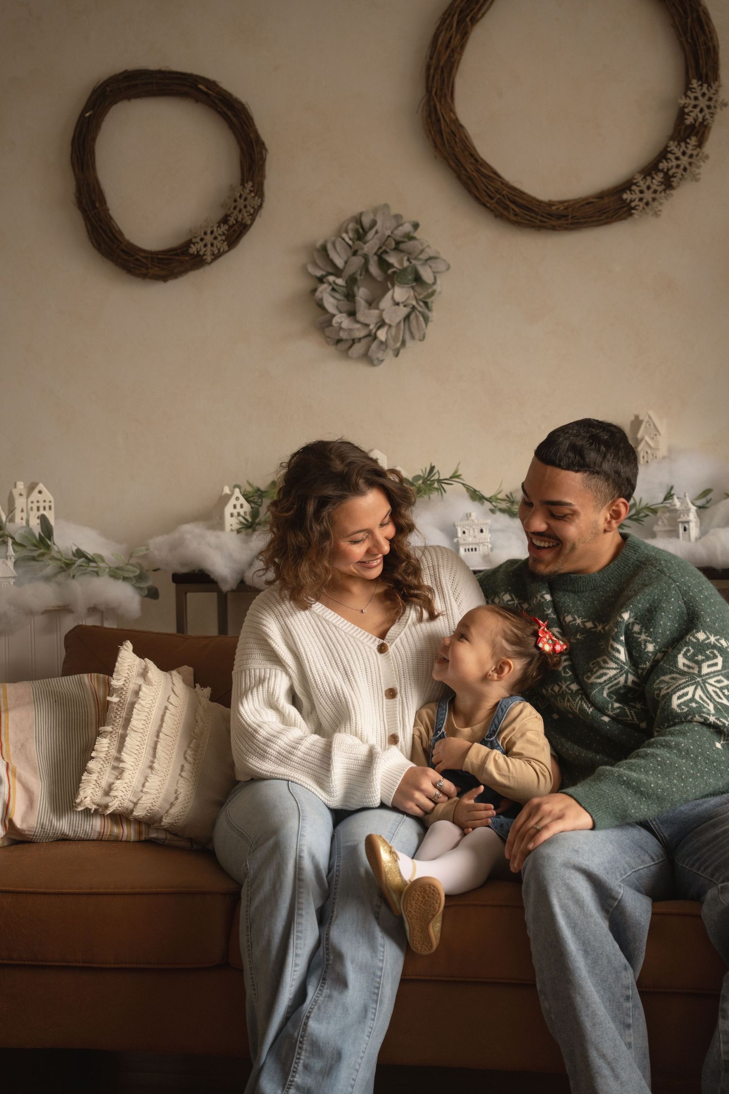 A couple in cozy sweaters share intimate moments while relaxing together on a brown couch in the studio with their child.