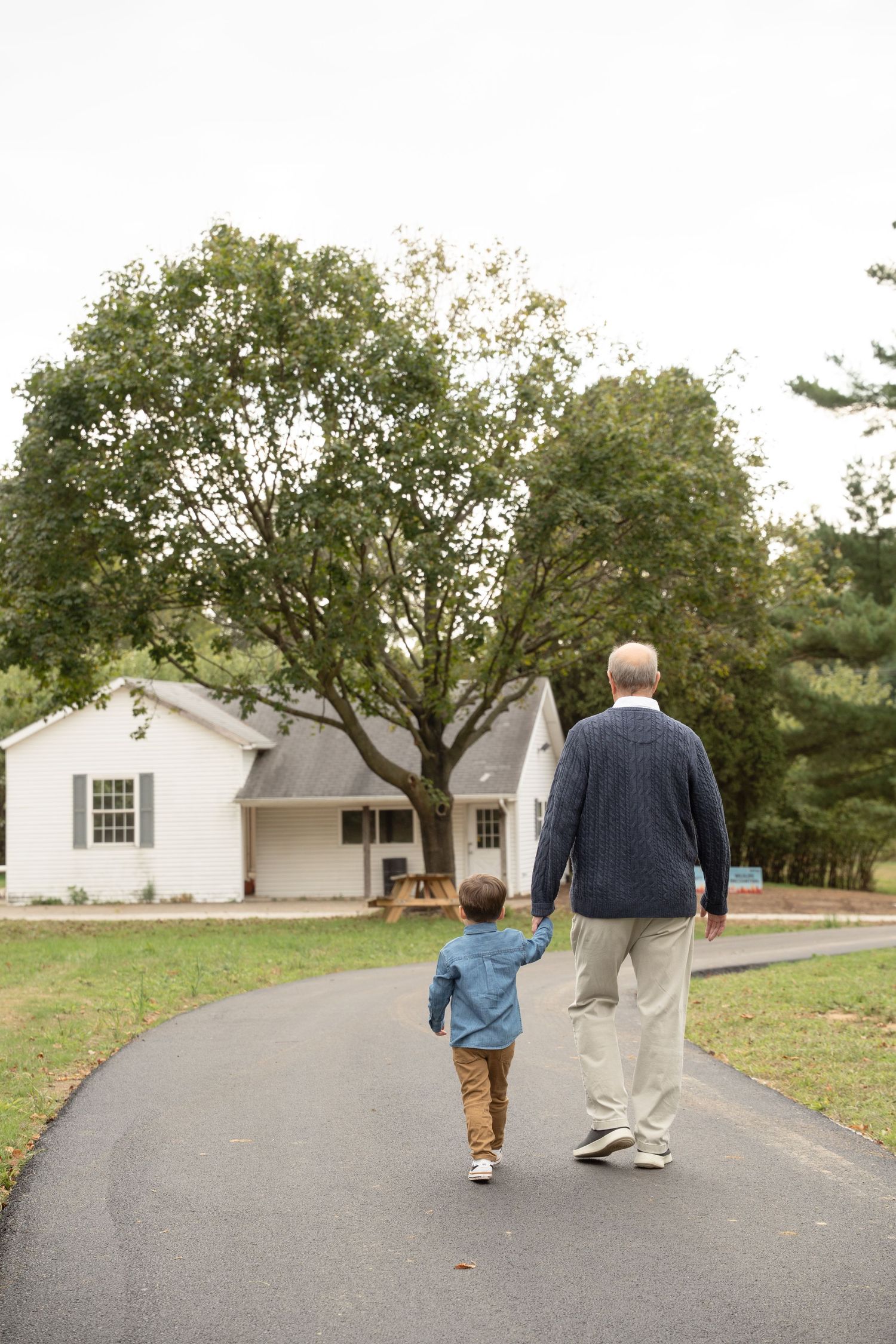 Little one with Grandpa walking together down a paved path surrounded by autumn trees and white buildings.
