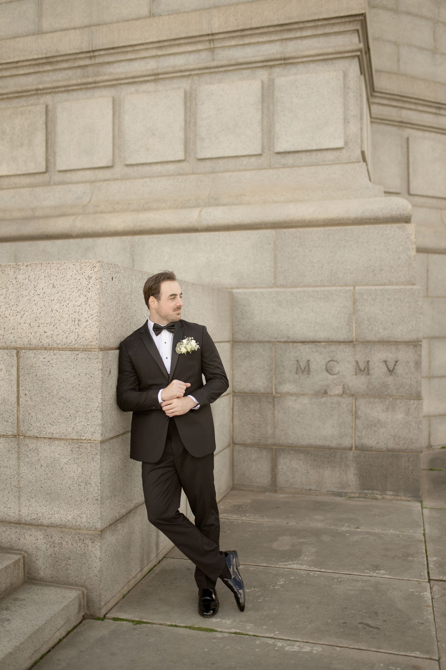 Groom in classic black tuxedo poses casually against stone building wall.