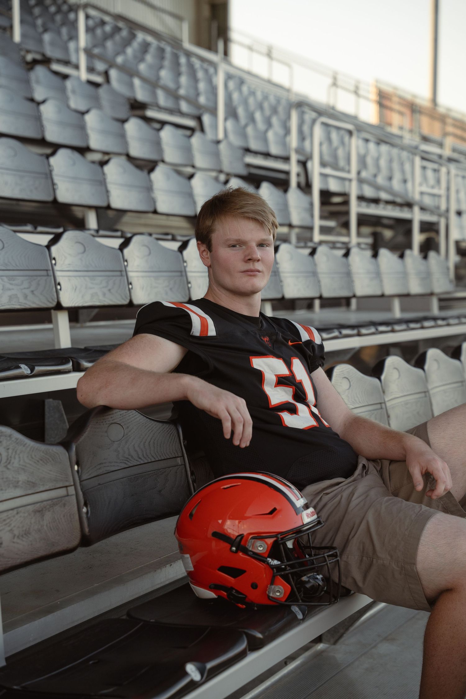 Football player reflects in stadium bleachers with football helmet during sports portrait session.