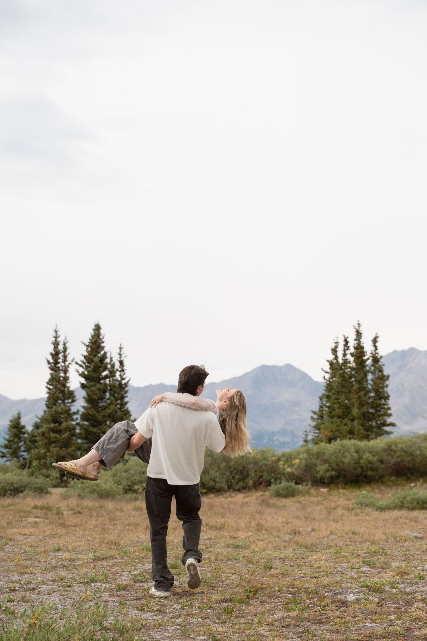 Couple dancing playfully in a Colorado mountain meadow with evergreen trees and peaks in the background.