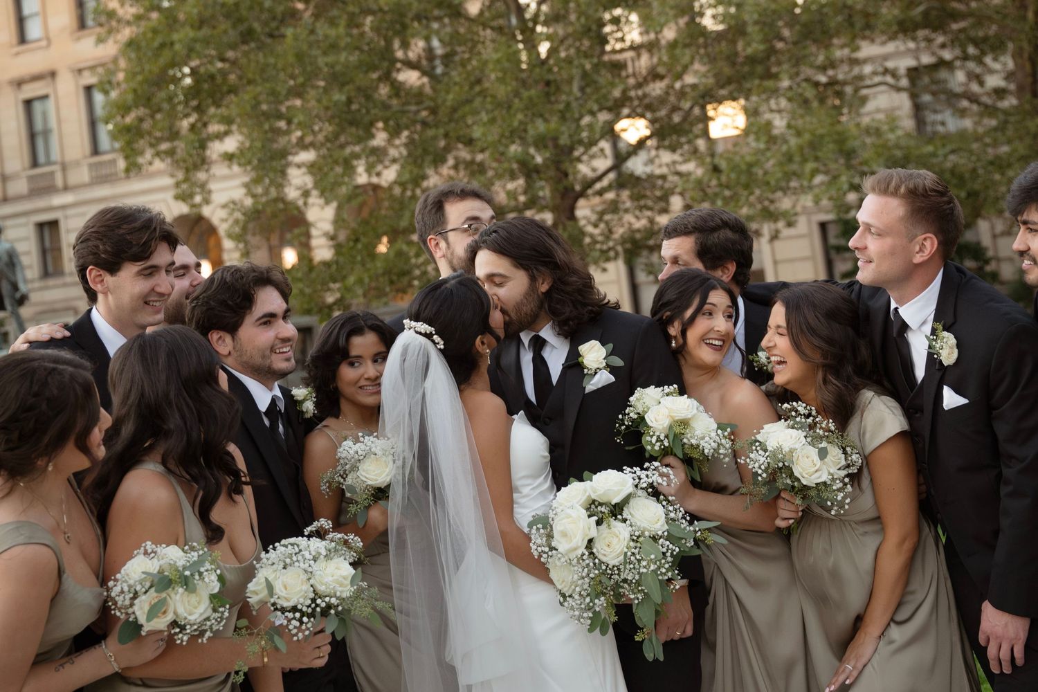 Wedding party members gather outdoors in formal attire with white floral bouquets near a stone building.