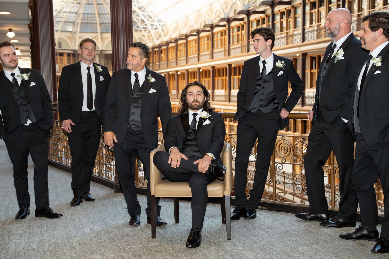A group of groomsmen in black tuxedos line up for a formal wedding photo in a rustic indoor venue.