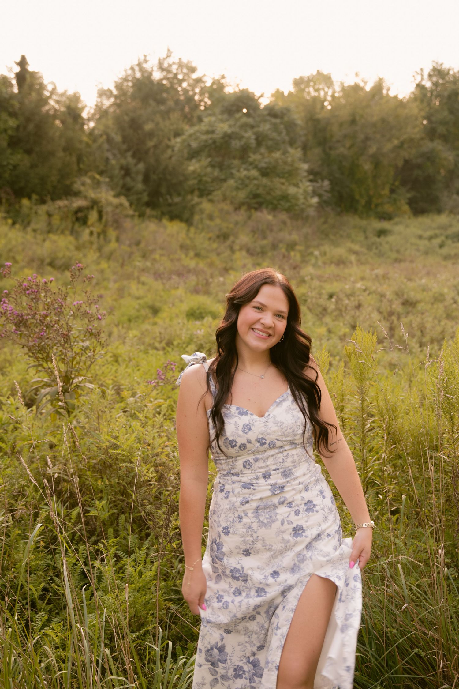 Senior in floral dress poses playfully in a sunlit meadow with tall grass.