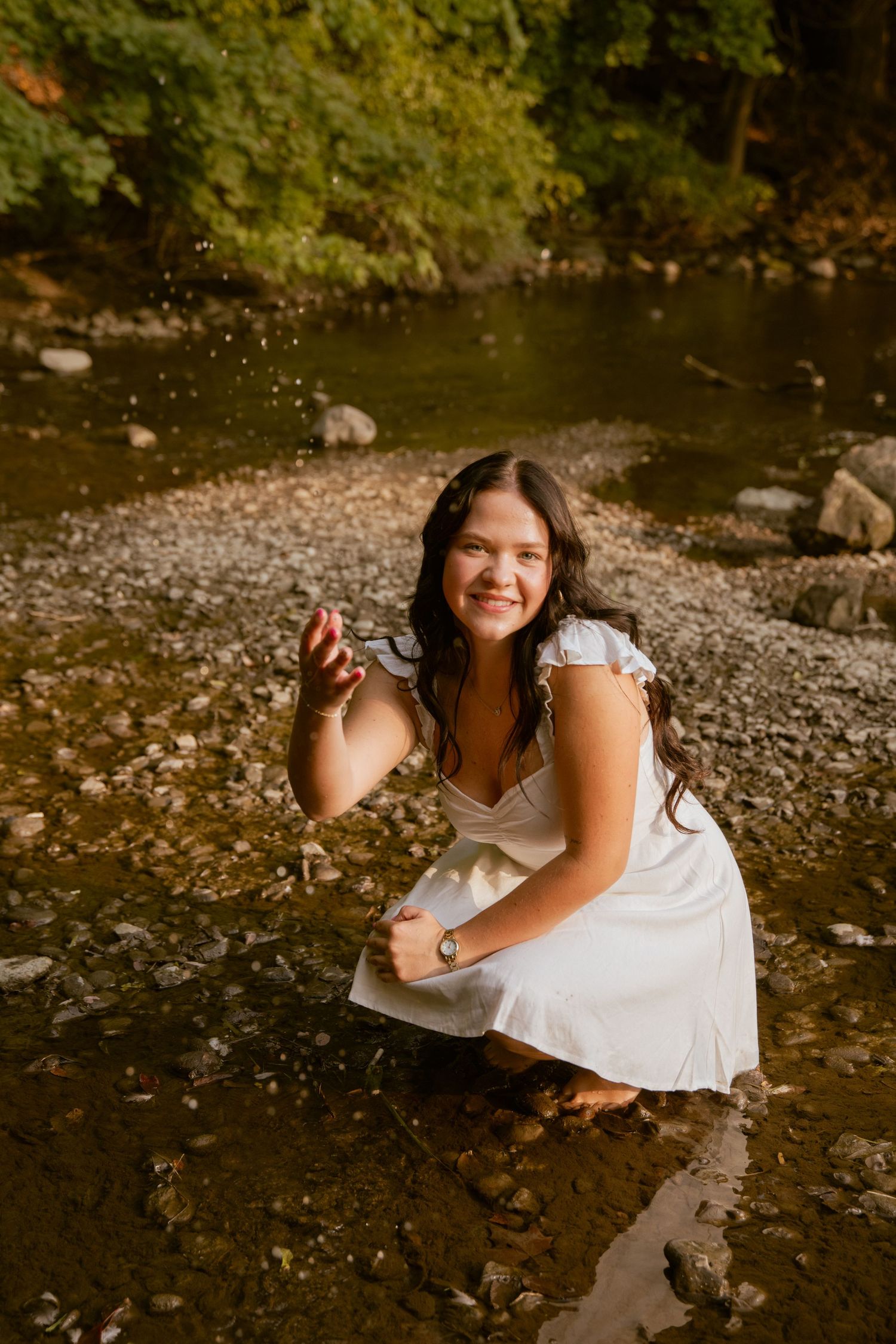 A senior in a white dress  by a rocky stream bank on a sunny day.