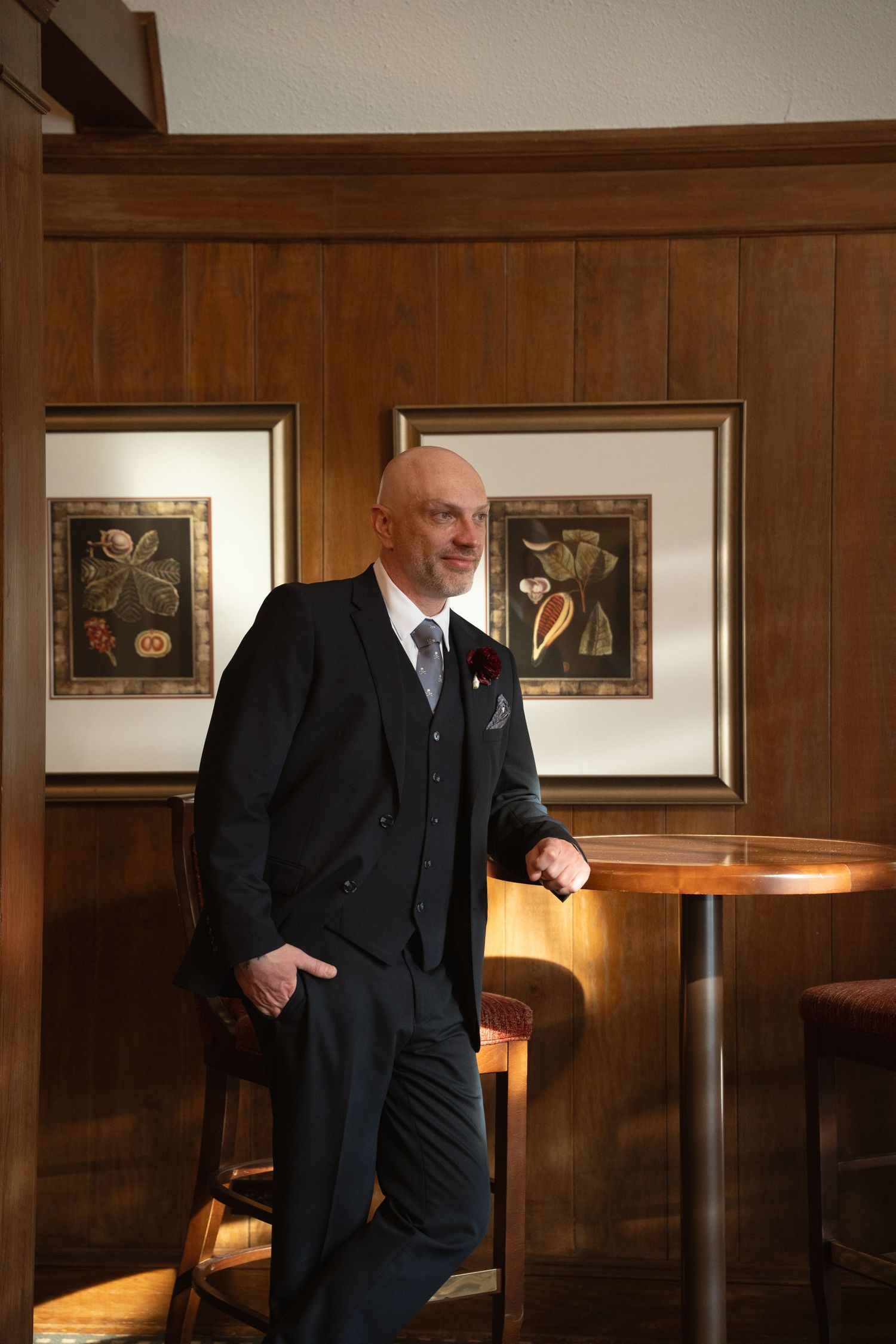 Elegant groom stands in wood-paneled room with framed artwork and warm ambient lighting.
