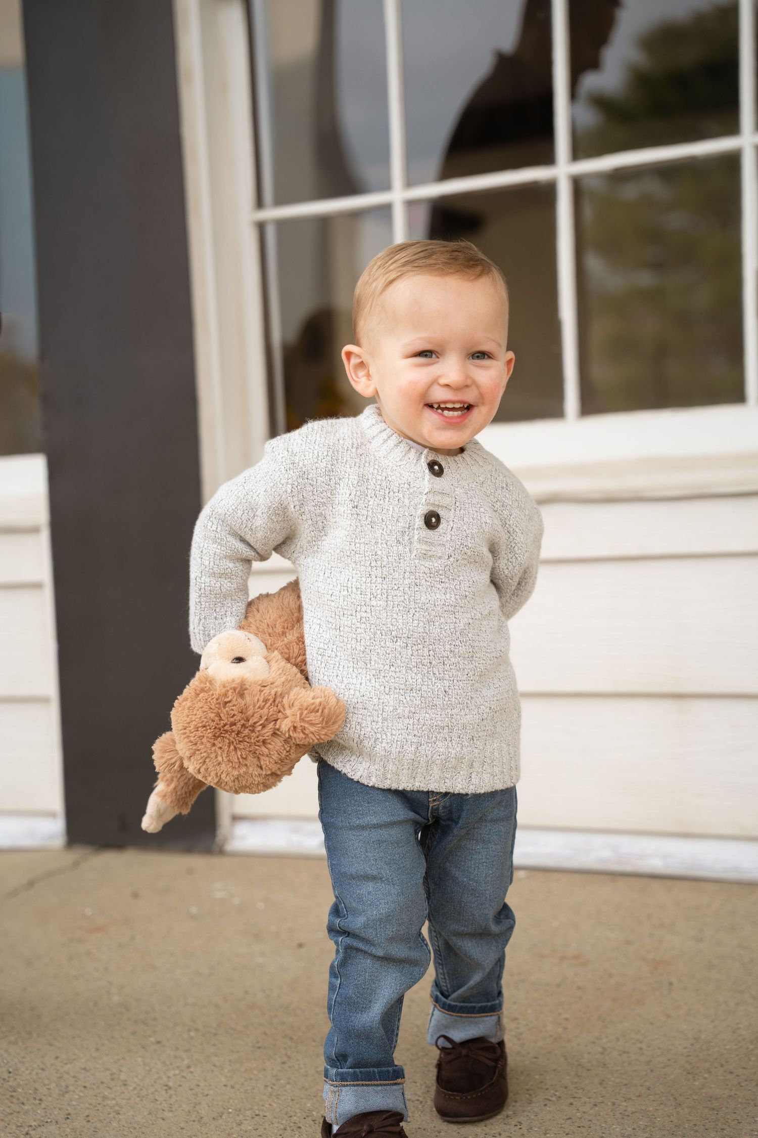 A little one in a gray sweater and jeans walks while holding a stuffed animal toy.