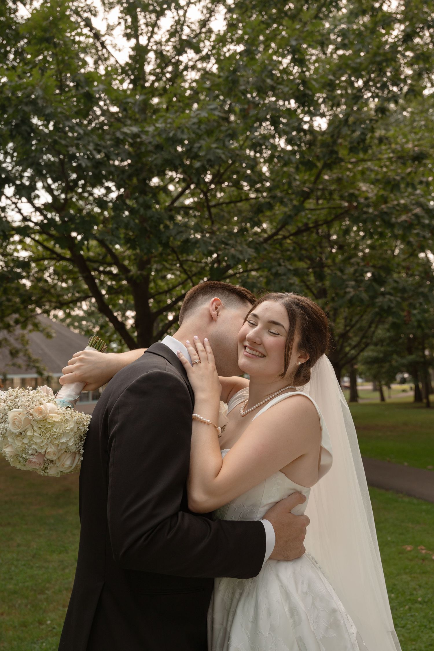 Newlyweds share a romantic moment outdoors among trees with white floral bouquet.