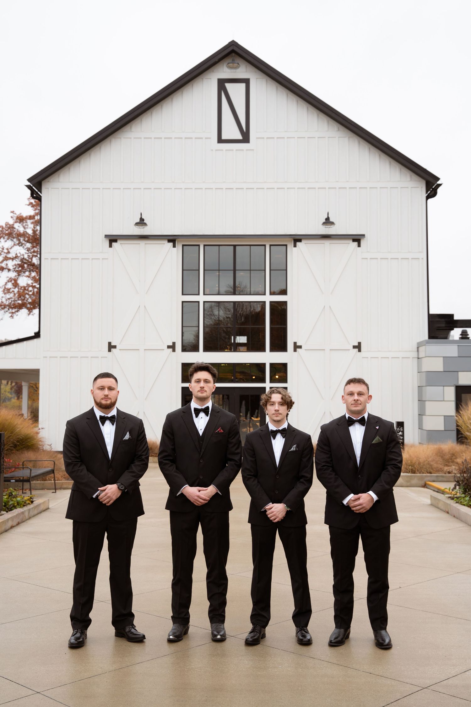 Groomsmen in black tuxedos pose sitting in line outside white barn venue.