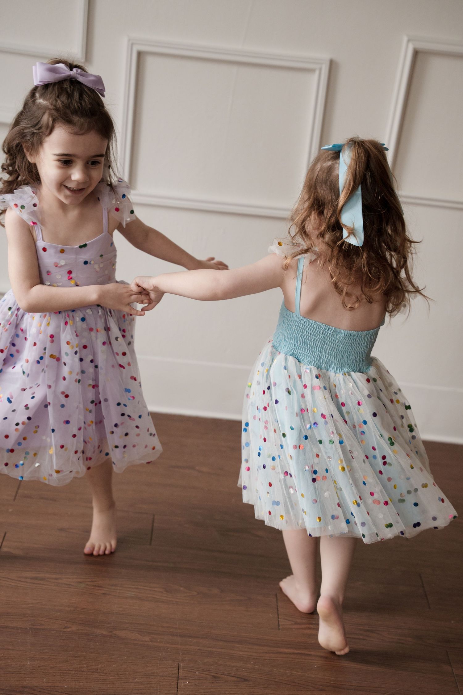 A child twirls in a sparkly pastel dress with polka dots on studio hardwood floors.