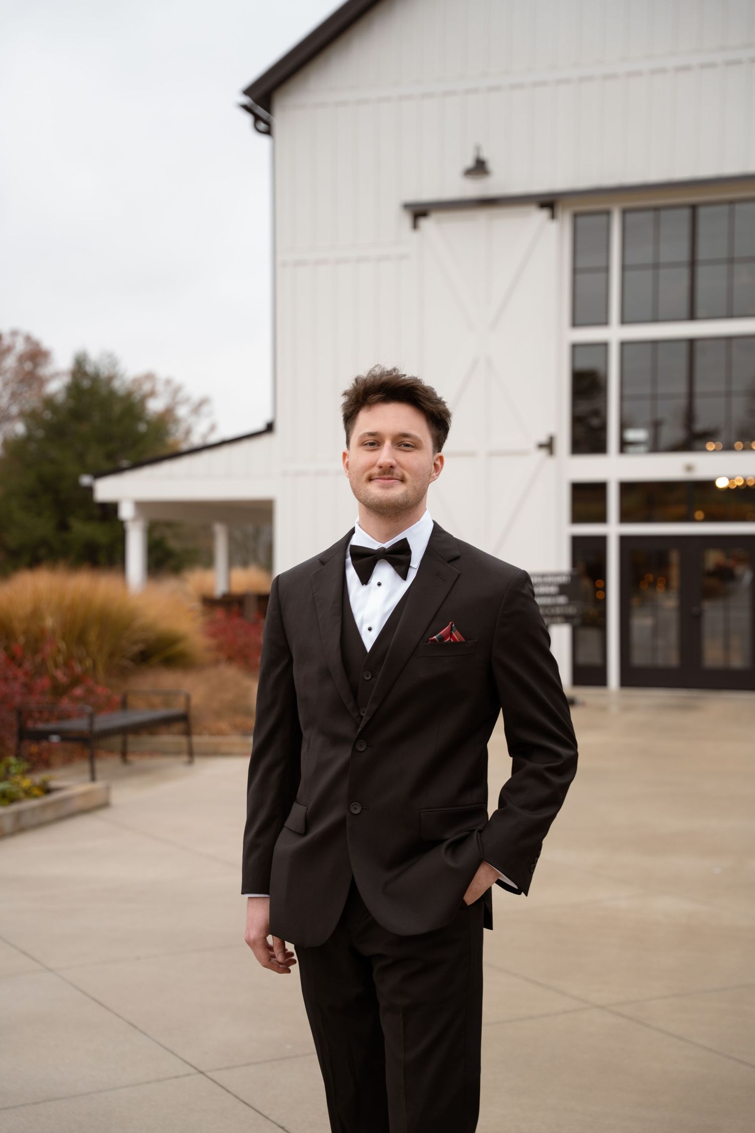 Groom in classic black tuxedo poses confidently outside white barn building.