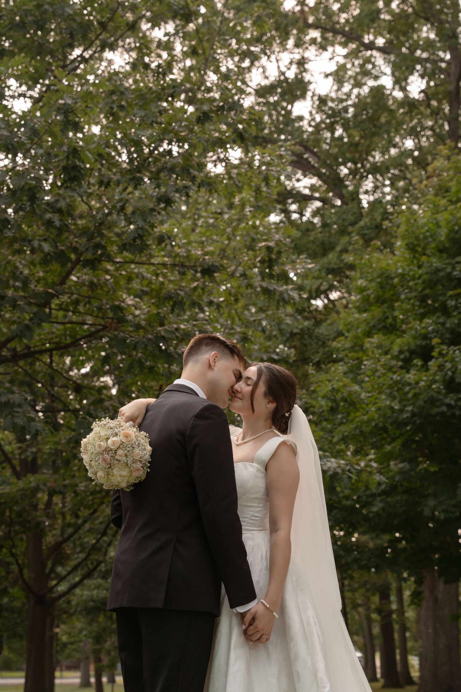 A romantic outdoor wedding moment with a couple embracing under trees while holding a white floral bouquet.