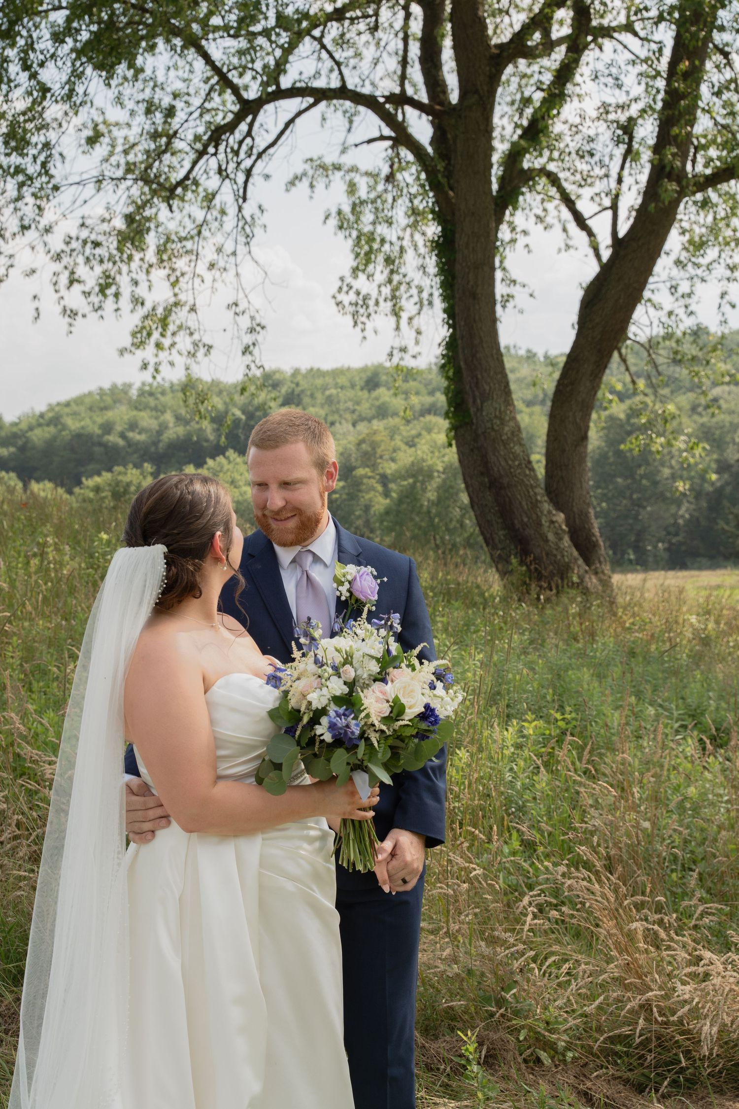 A wedding couple stands together in a Pennsylvania meadow holding bouquet of purple and white flowers.