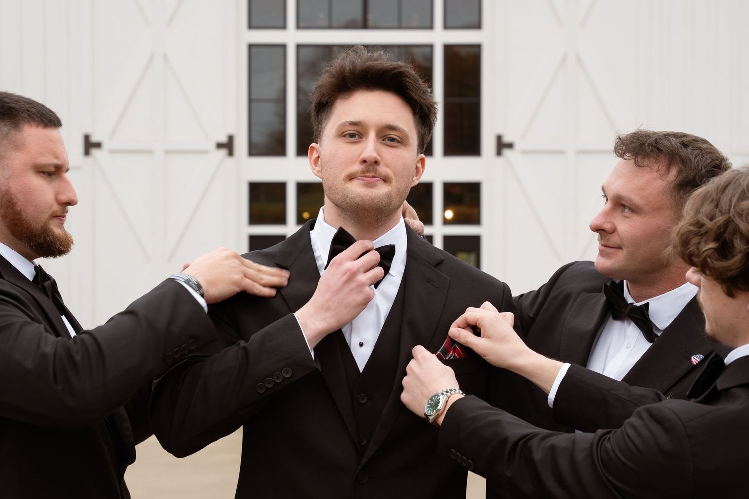 Groomsmen in black suits share a lighthearted moment adjusting grooms attire outside a wedding venue