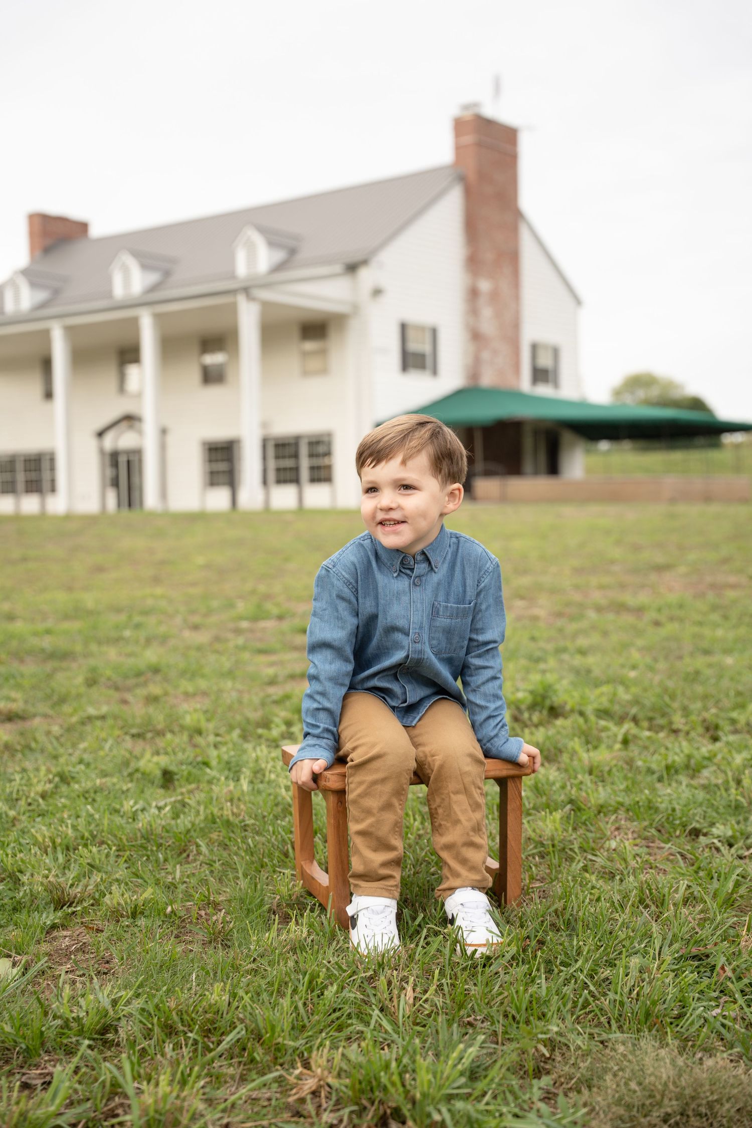 Little one sits in wooden chair on grassy hill with white farmhouse buildings in background.