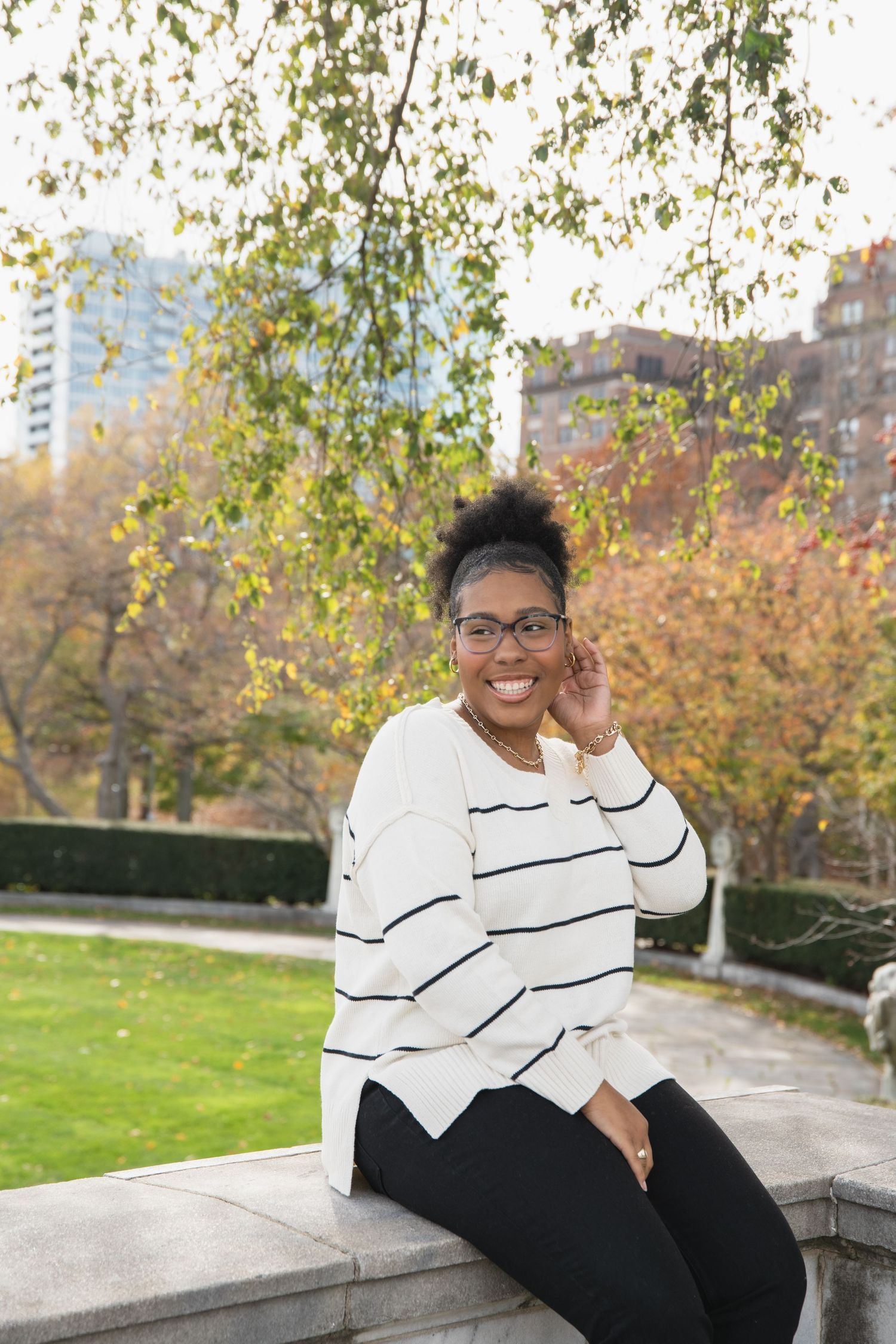 A senior in a black and white striped sweater sits on a stone ledge surrounded by autumn foliage.