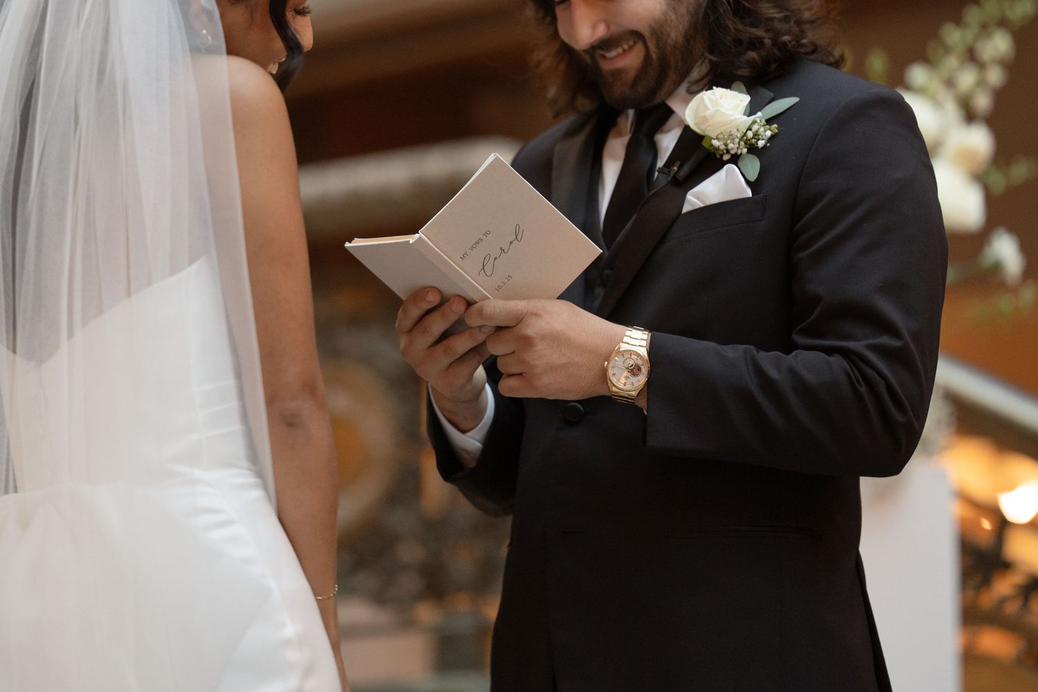 Groom in black suit reads wedding vows from a small book during an intimate ceremony.