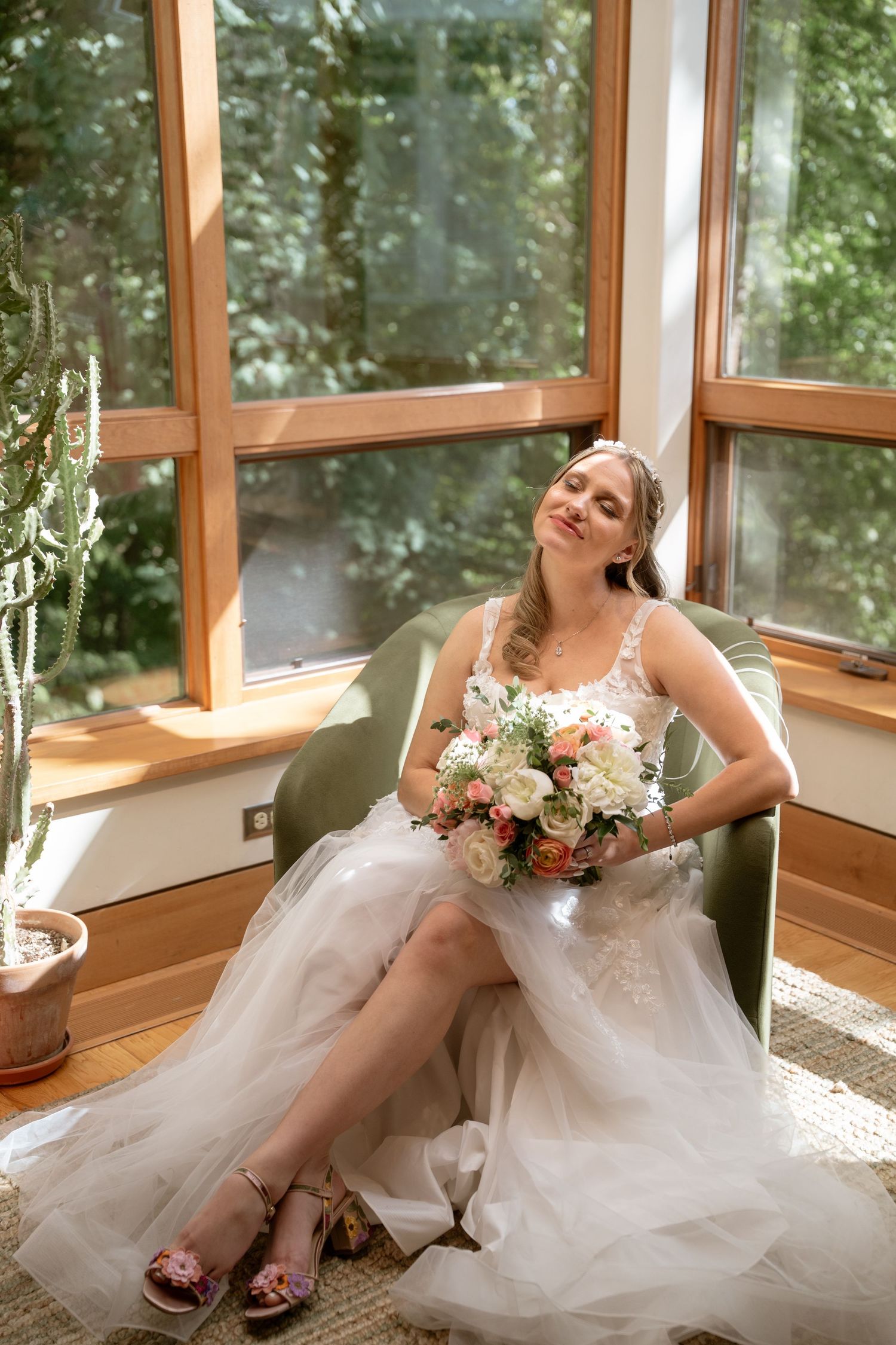 Bride in white gown relaxes in sunlit room with wooden window frames.