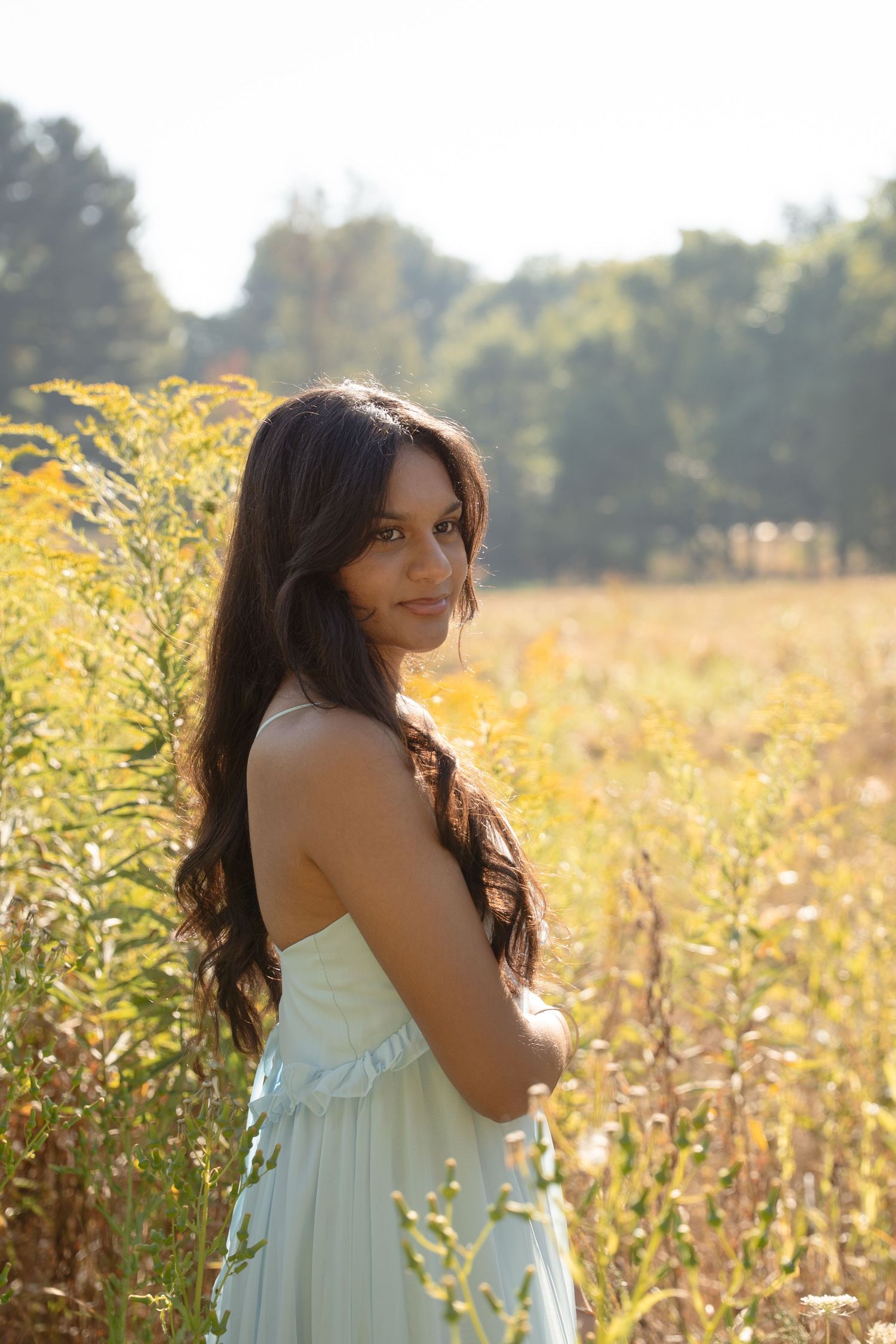 Senior in flowing white dress poses gracefully in sunlit field of golden wildflowers.