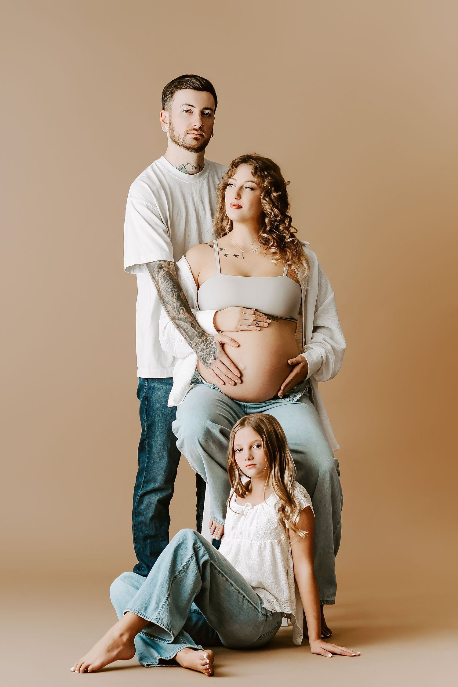 A family maternity photoshoot featuring warm neutral tones and casual attire against a beige backdrop.