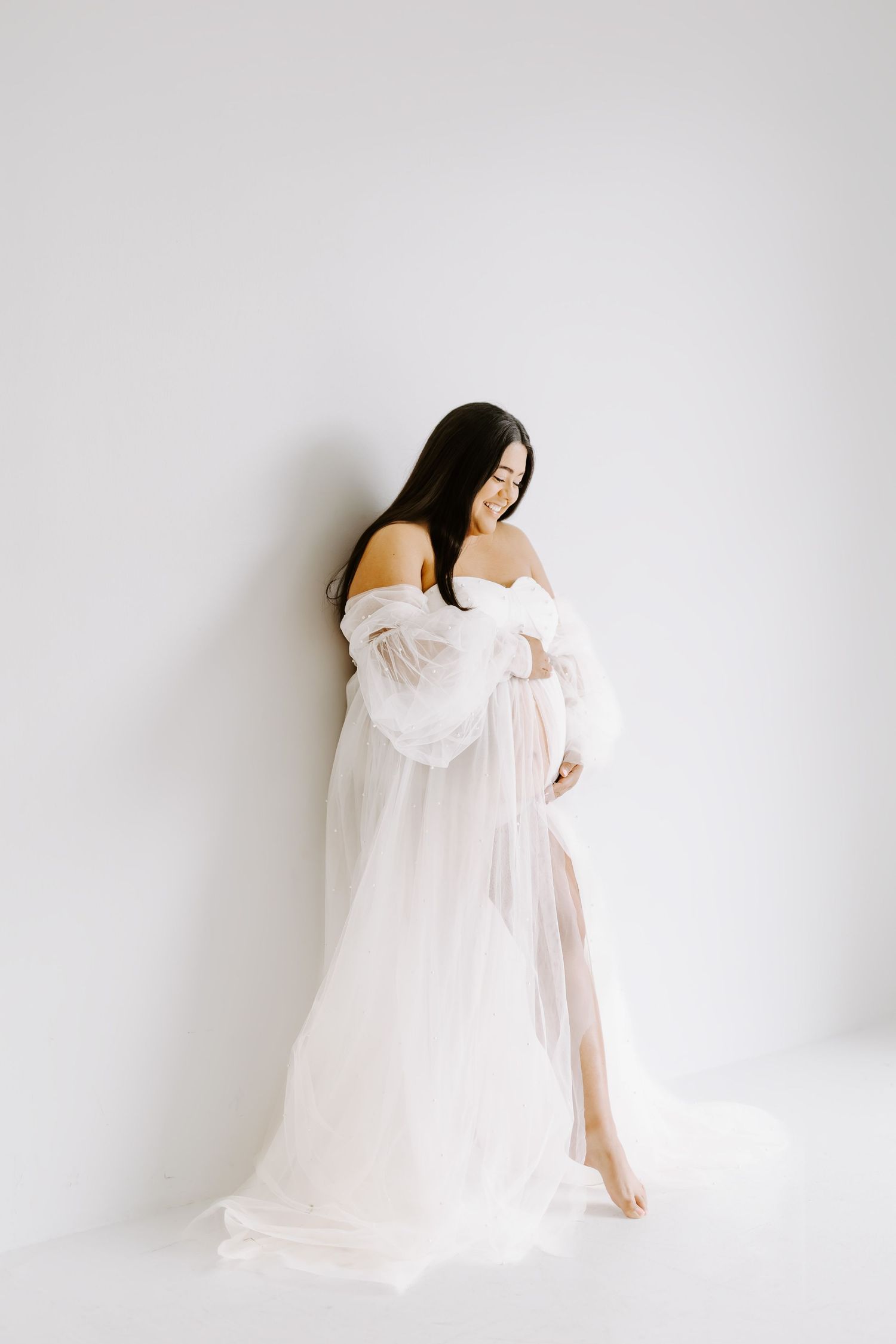 Ethereal maternity portraits with woman in flowing white dress against white backdrop.