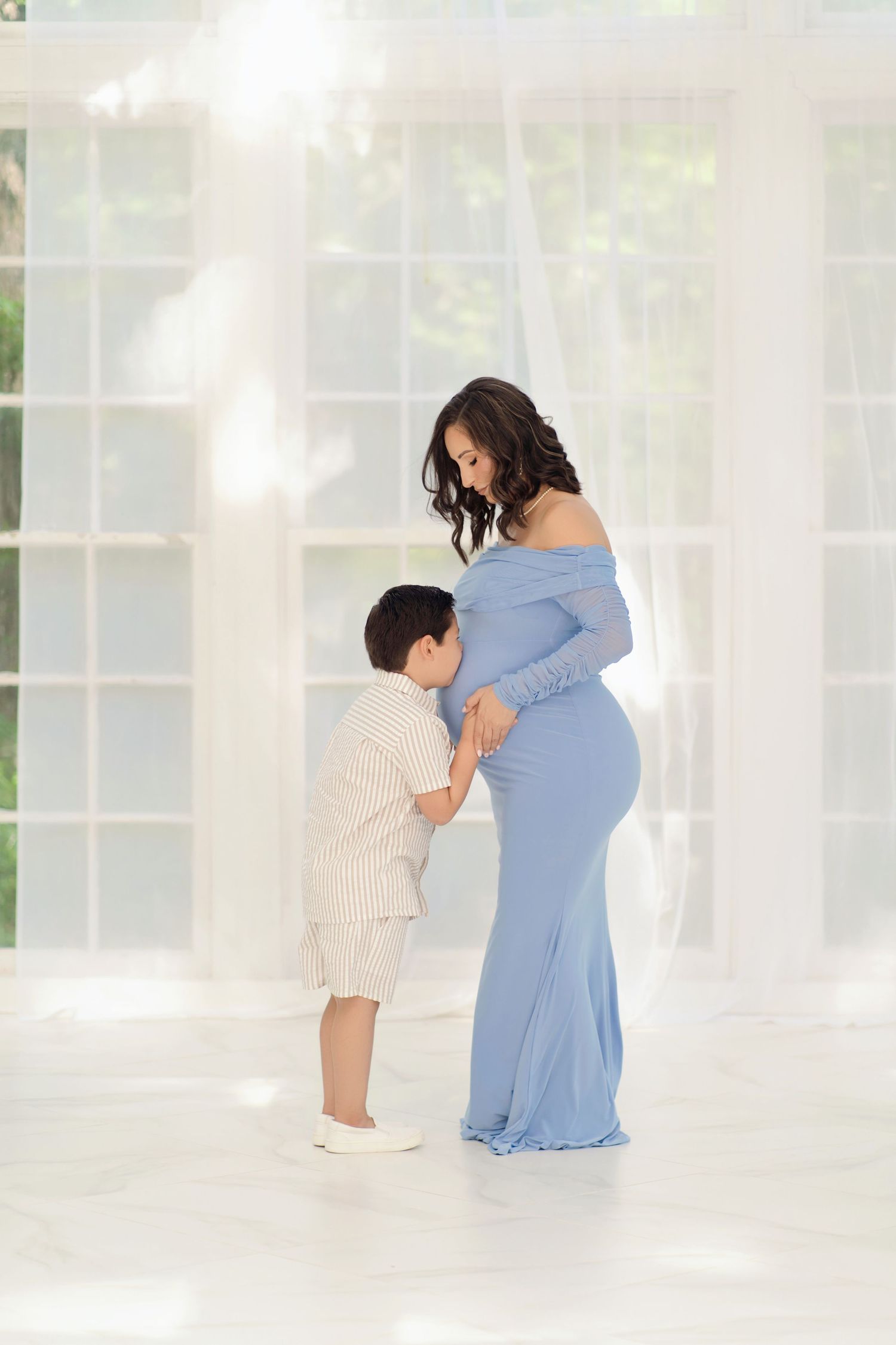 A pregnant woman in a blue gown shares a tender moment with her young son in a bright, airy white room.